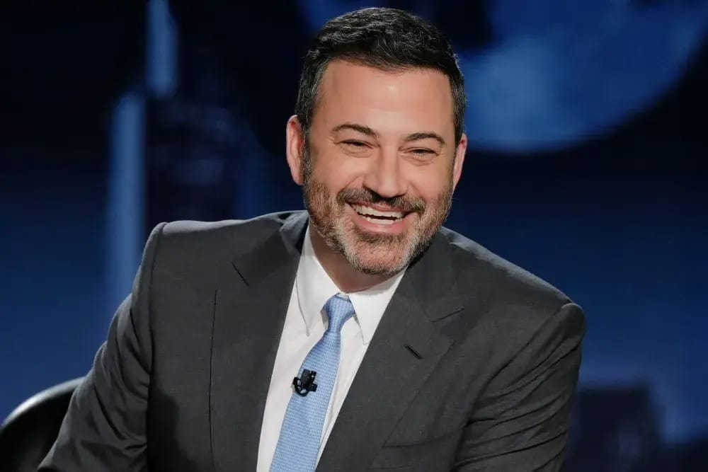 A man in a suit and tie smiles while sitting in a studio, reminiscent of ABC’s Jimmy Kimmel Live set.