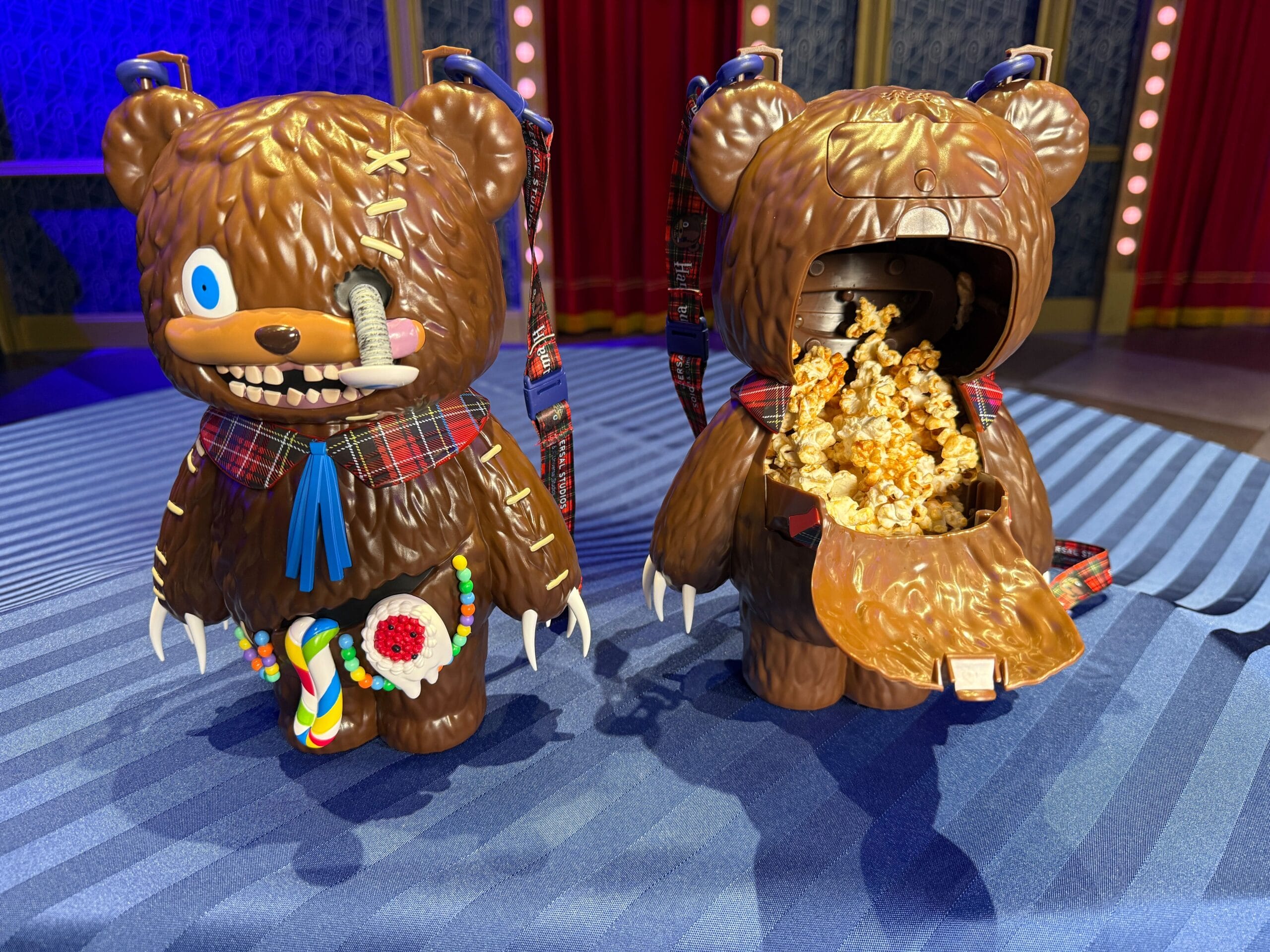 Two bear-shaped popcorn buckets with quirky designs, one open to reveal popcorn inside, displayed on a blue-striped table.