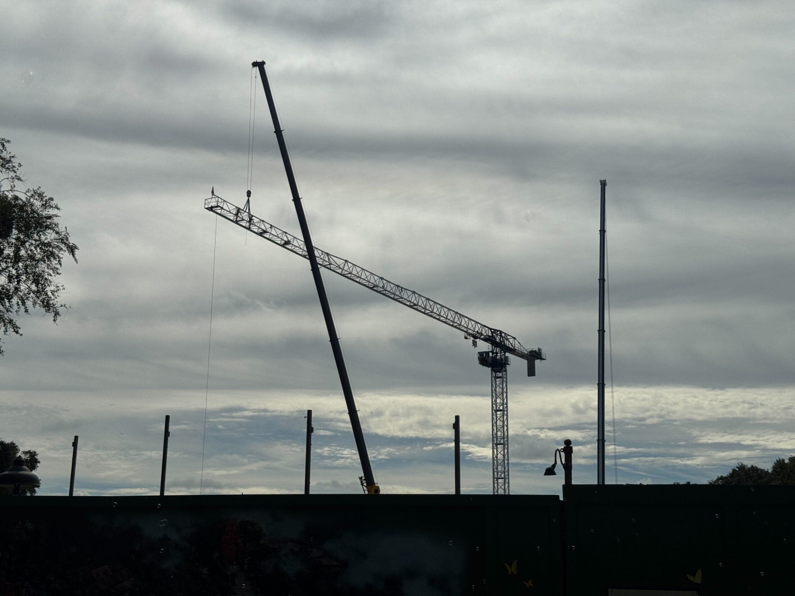 A Tower Crane and construction equipment at a work site under a cloudy sky, fencing in the foreground.