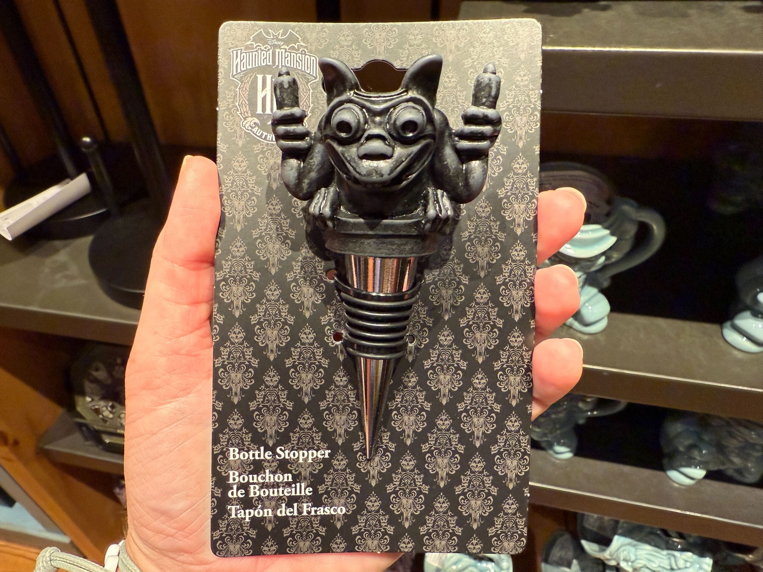 A hand holds a Haunted Mansion-themed bottle stopper featuring a small gargoyle figurine with raised arms.