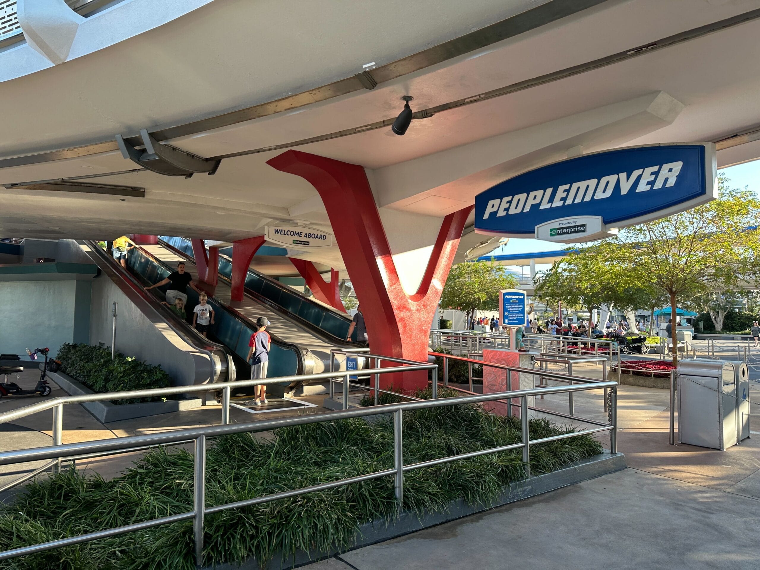 Two escalators lead to the PeopleMover ride entrance under a white canopy with blue and red accents at a theme park.
