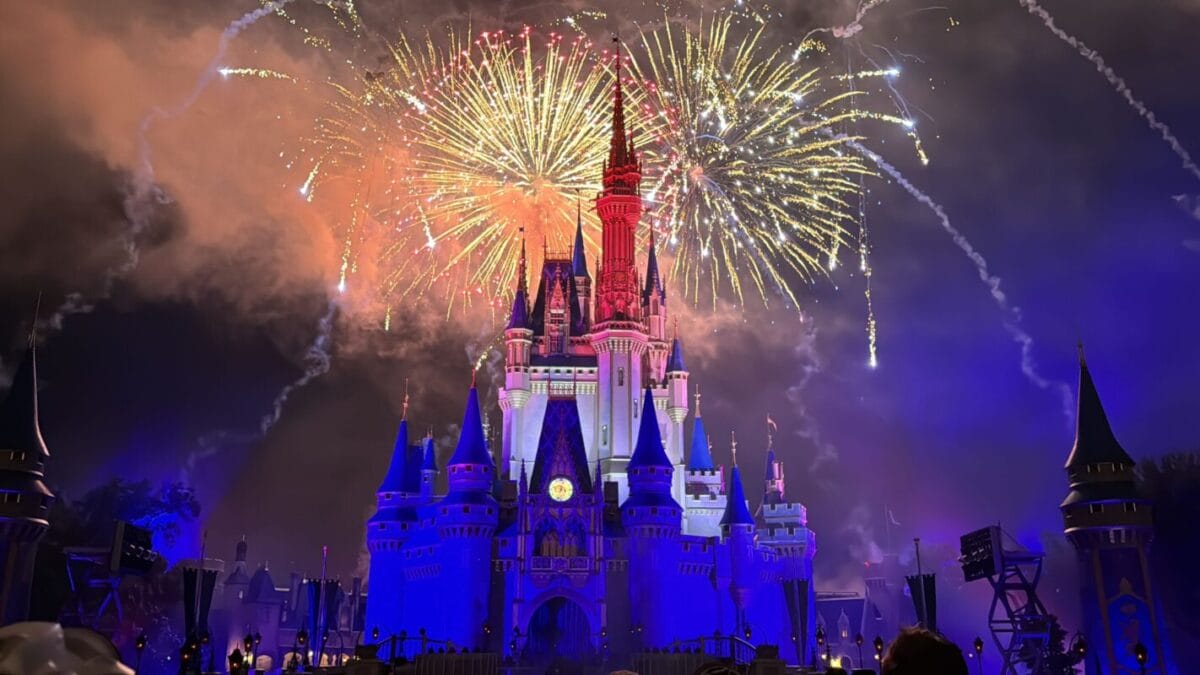 Disney fireworks burst over a castle resembling Magic Kingdom's, its base aglow in rainbow lights, dazzling the 2026 night sky.