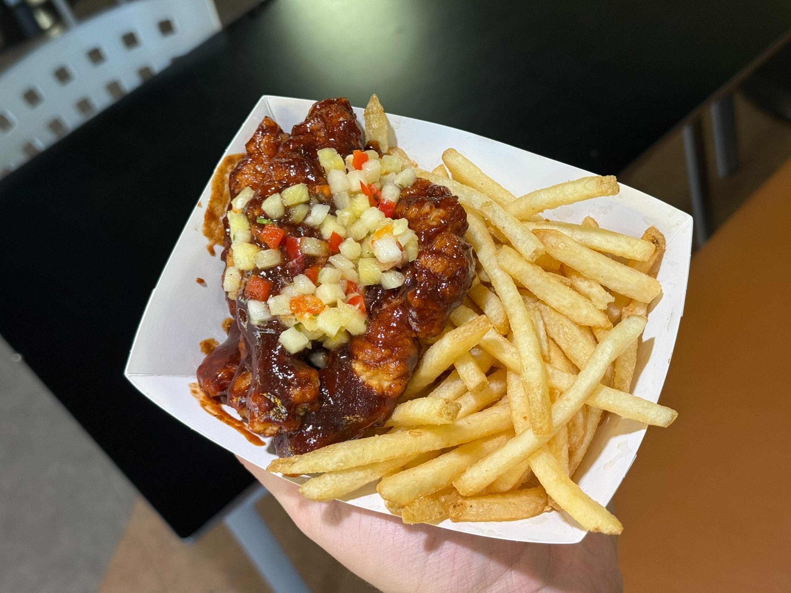 A hand holds a Disney Parks food tray filled with crispy Chicken Strips and saucy fried chicken topped with colorful veggies.