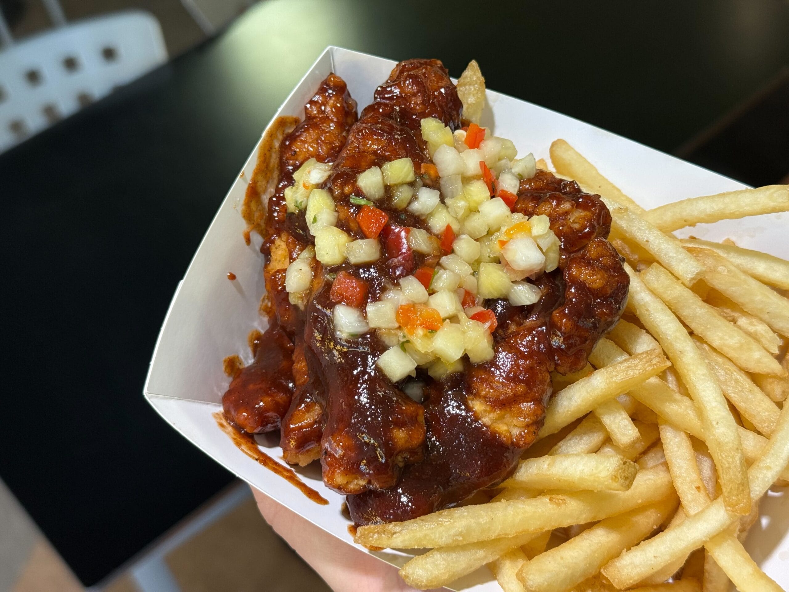 A hand holds a Disney Parks food tray filled with Jerk BBQ chicken strips topped with pineapple salsa and served with fries.