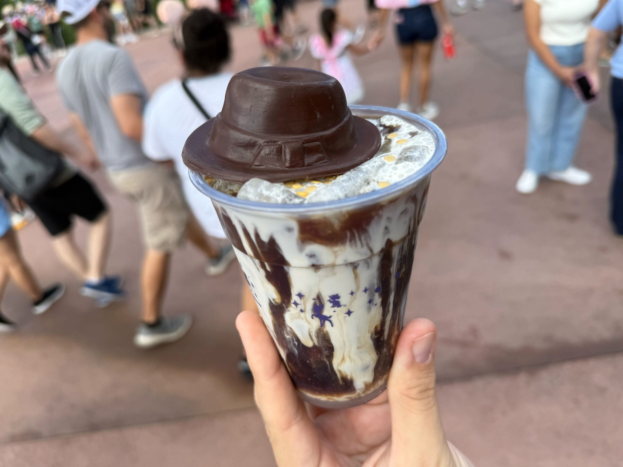 A hand holds an Epic Cold Brew dessert with chocolate drizzle at Disney's Hollywood Studios, surrounded by park guests and themed decor.