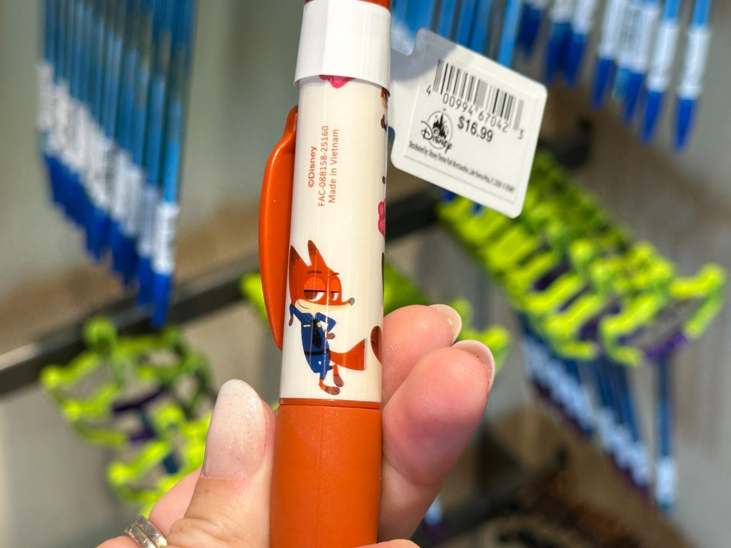 A hand at a Disney park holds an orange Zootopia pen with Nick Wilde; other pens hang on a display, typical of park shops.