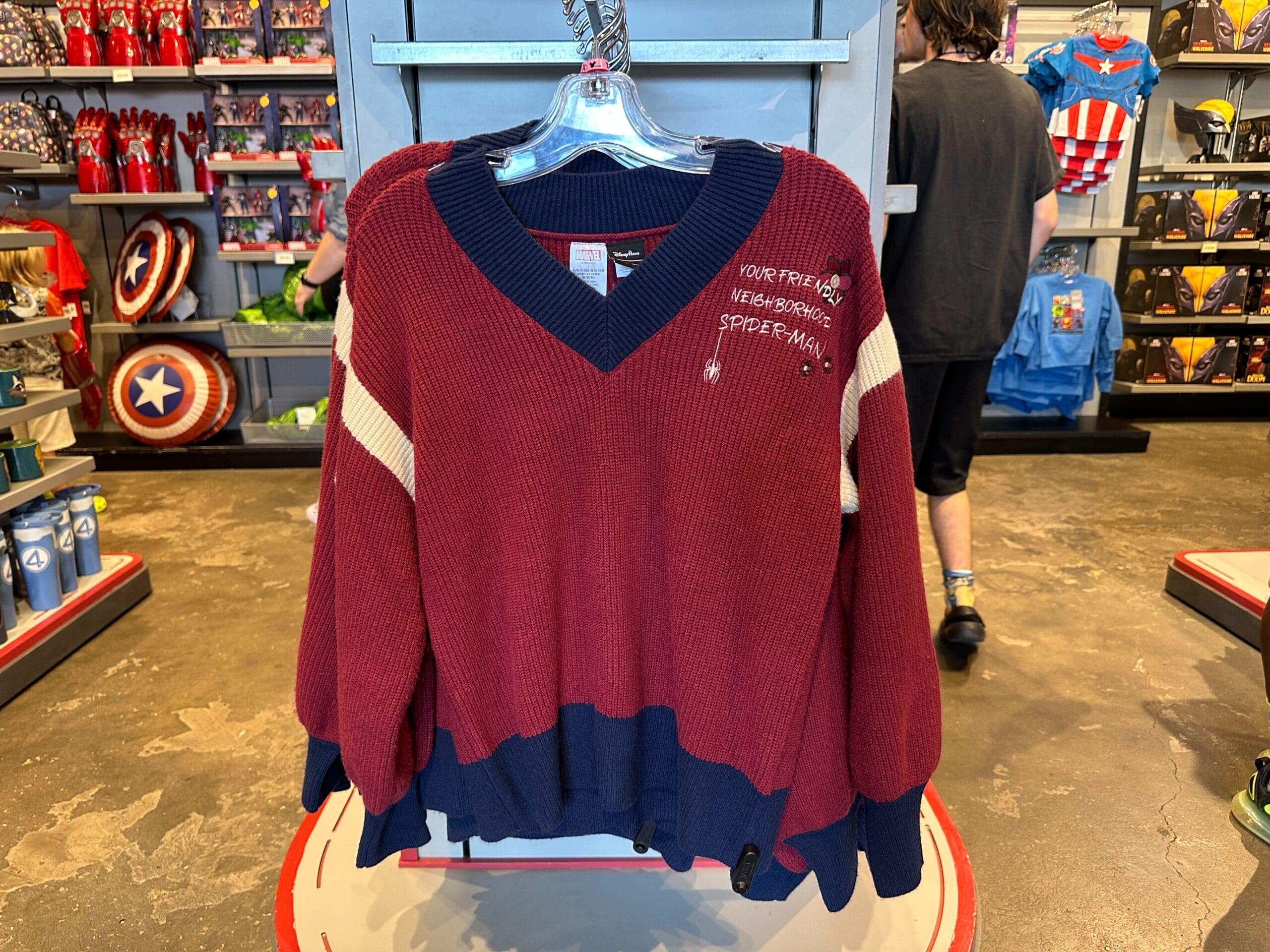 A red and navy Spider-Man sweater by Her Universe with cream stripes hangs among Marvel merchandise at a Disney park shop.