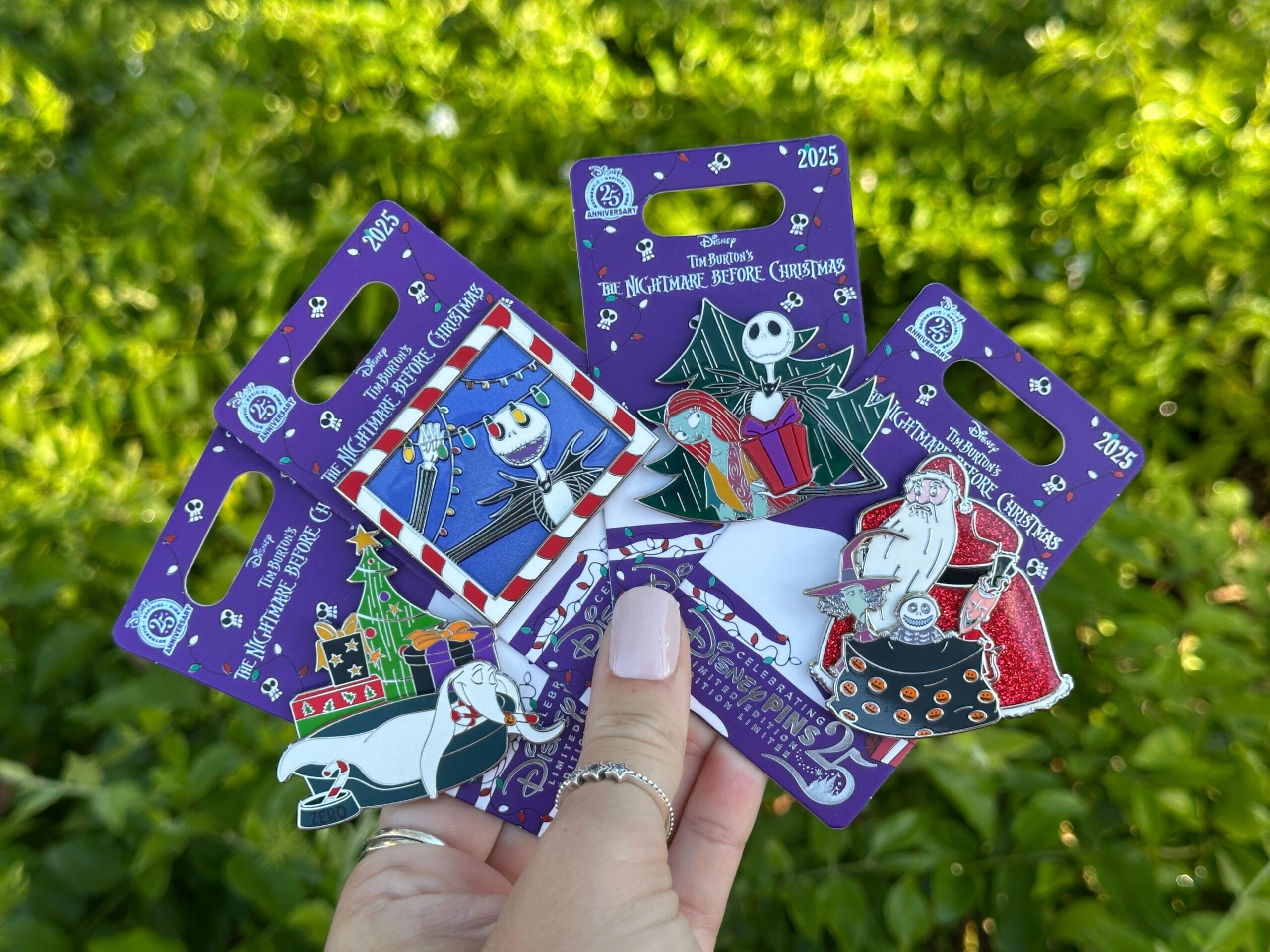 A hand displays four Nightmare Before Christmas pins from Disney World, held up before lush greenery—no castle in view.
