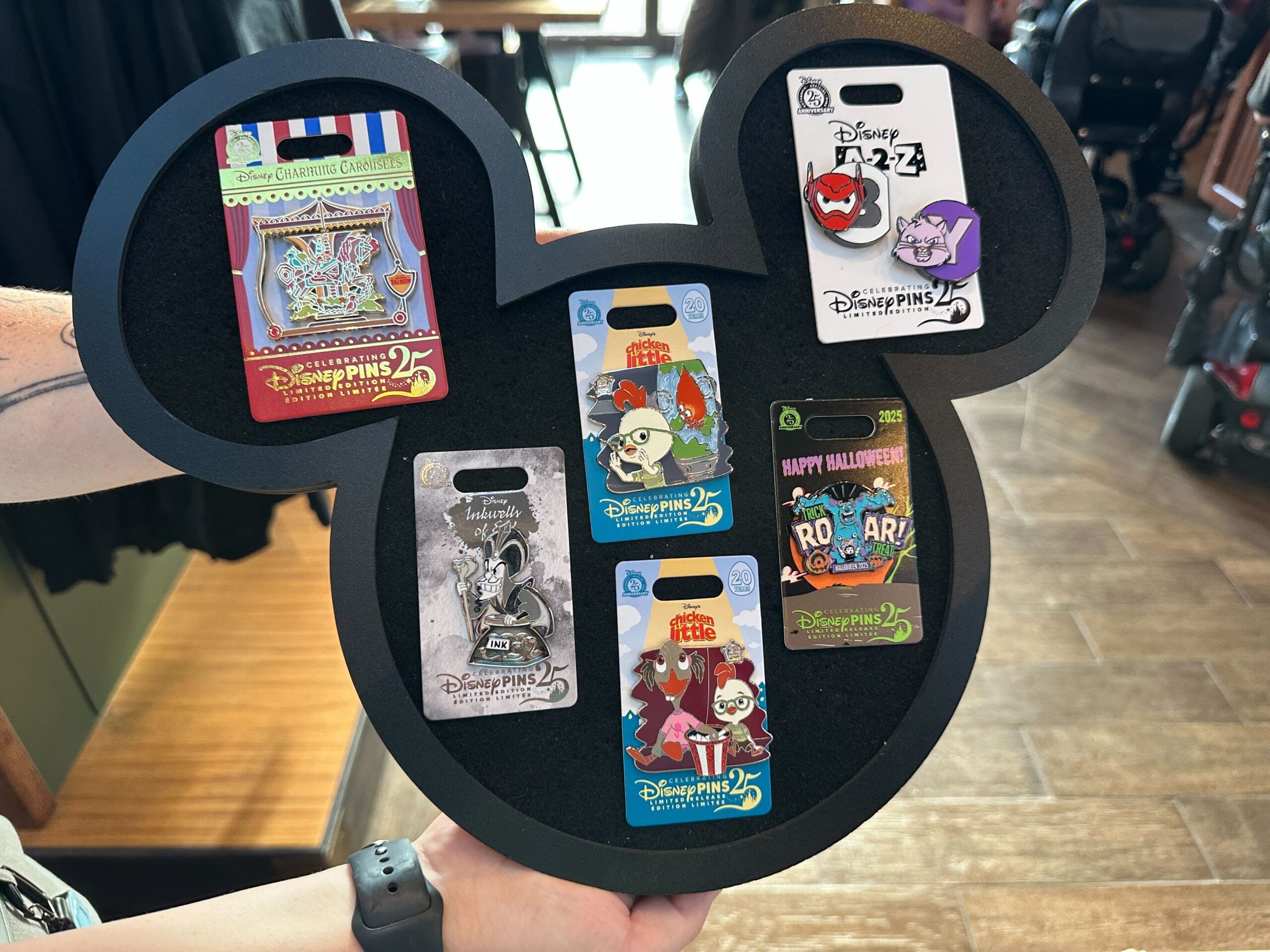 A person displays a Mickey Mouse-shaped pin board holding six Disney Park pins, including a Tiki-Themed Carousel design. No castles seen.