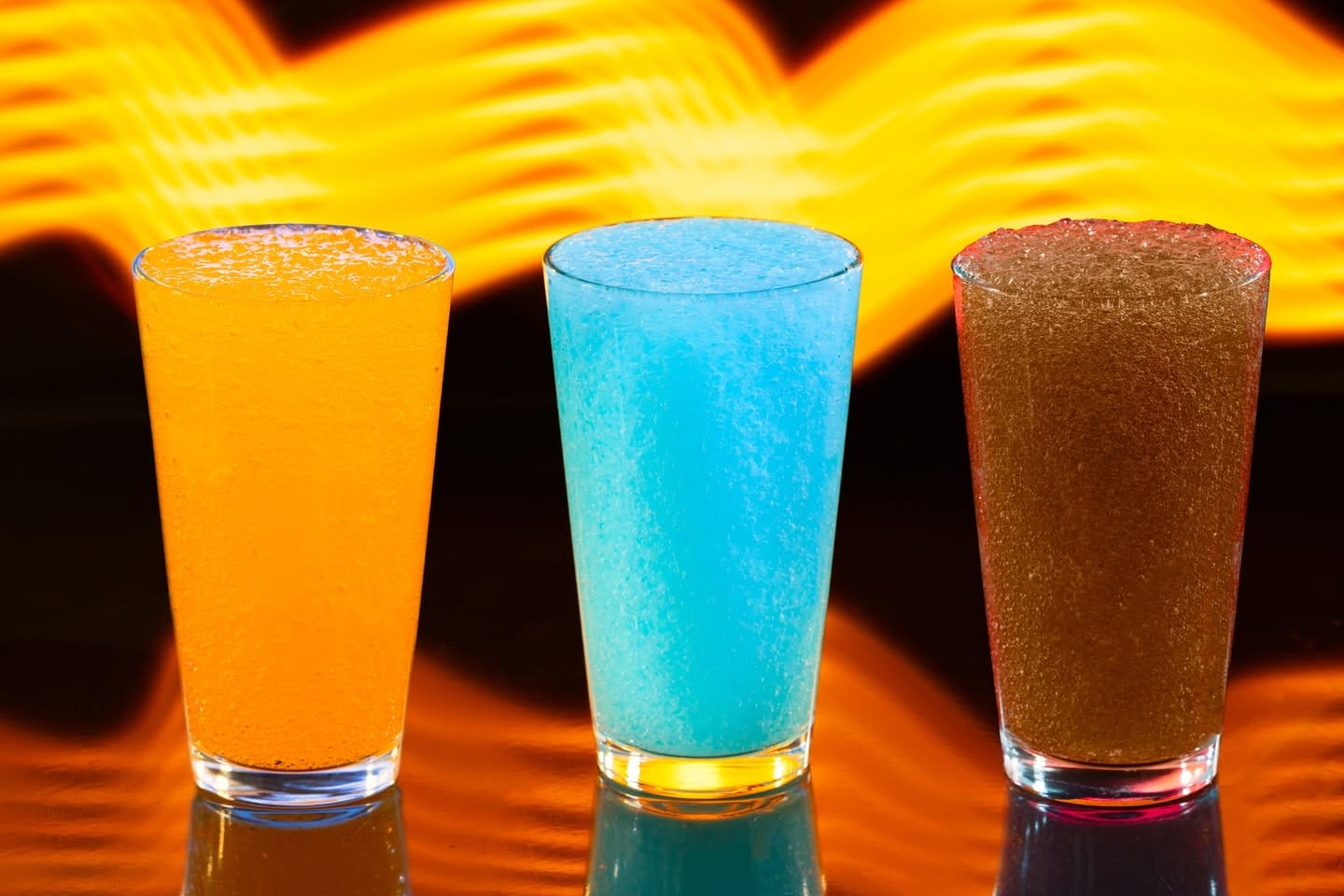 Three Astro Fizz slushy drinks lined up in front of a bright, wavy orange background, reminiscent of Tomorrowland at Disney Parks.