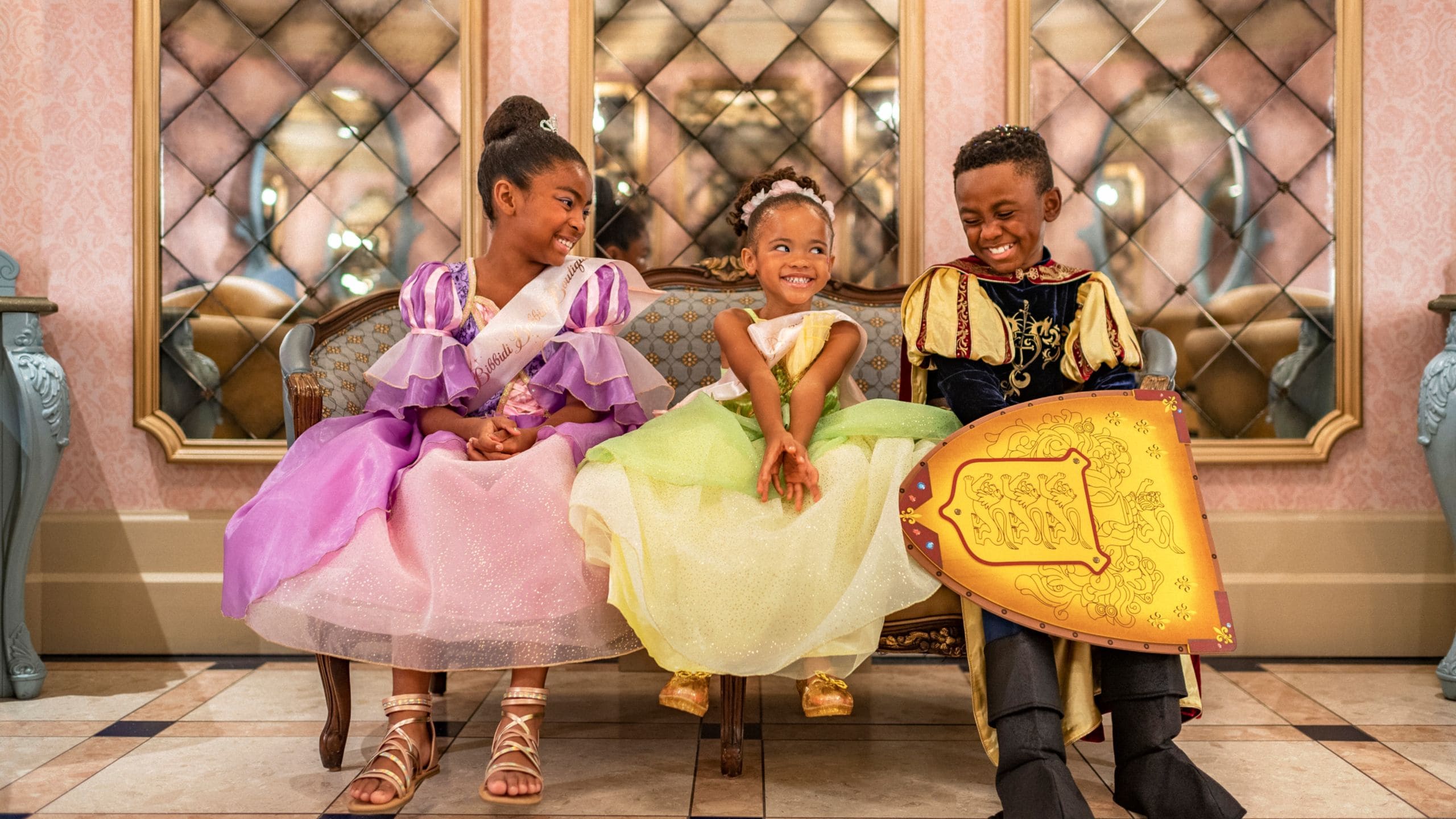 Three smiling kids in princess and knight costumes sit on a bench, echoing the royal flair of a Disney Park’s castle interior.