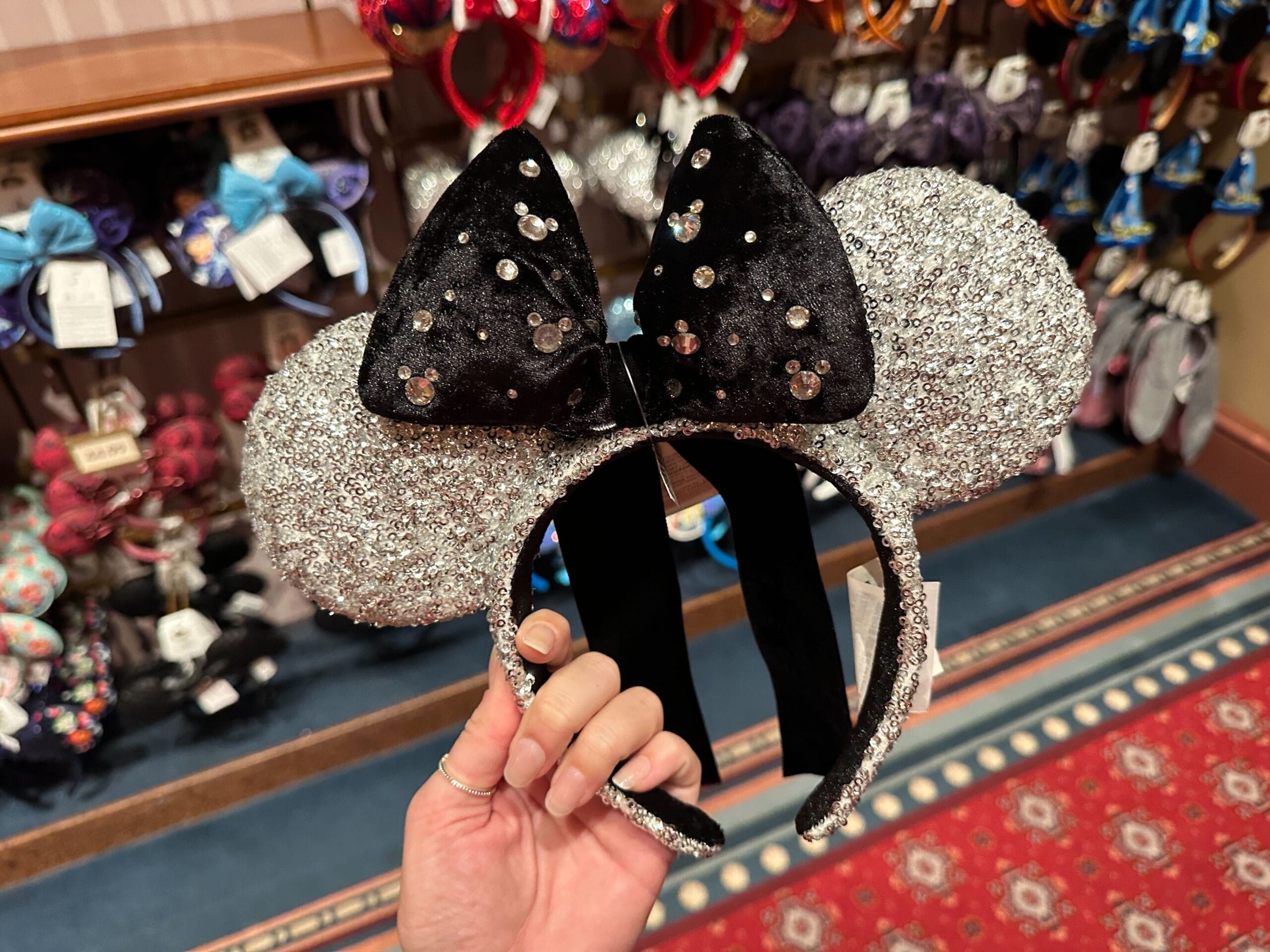 A hand displays a sparkly silver Minnie Mouse ear headband with a black sequined bow, likely in a Disney park merchandise shop.