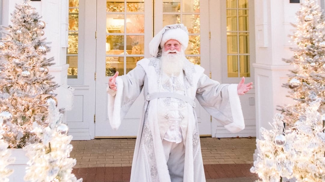 A man in white Santa robes stands between snowy, decorated trees outside Disney’s BoardWalk, with no castle visible in the scene.