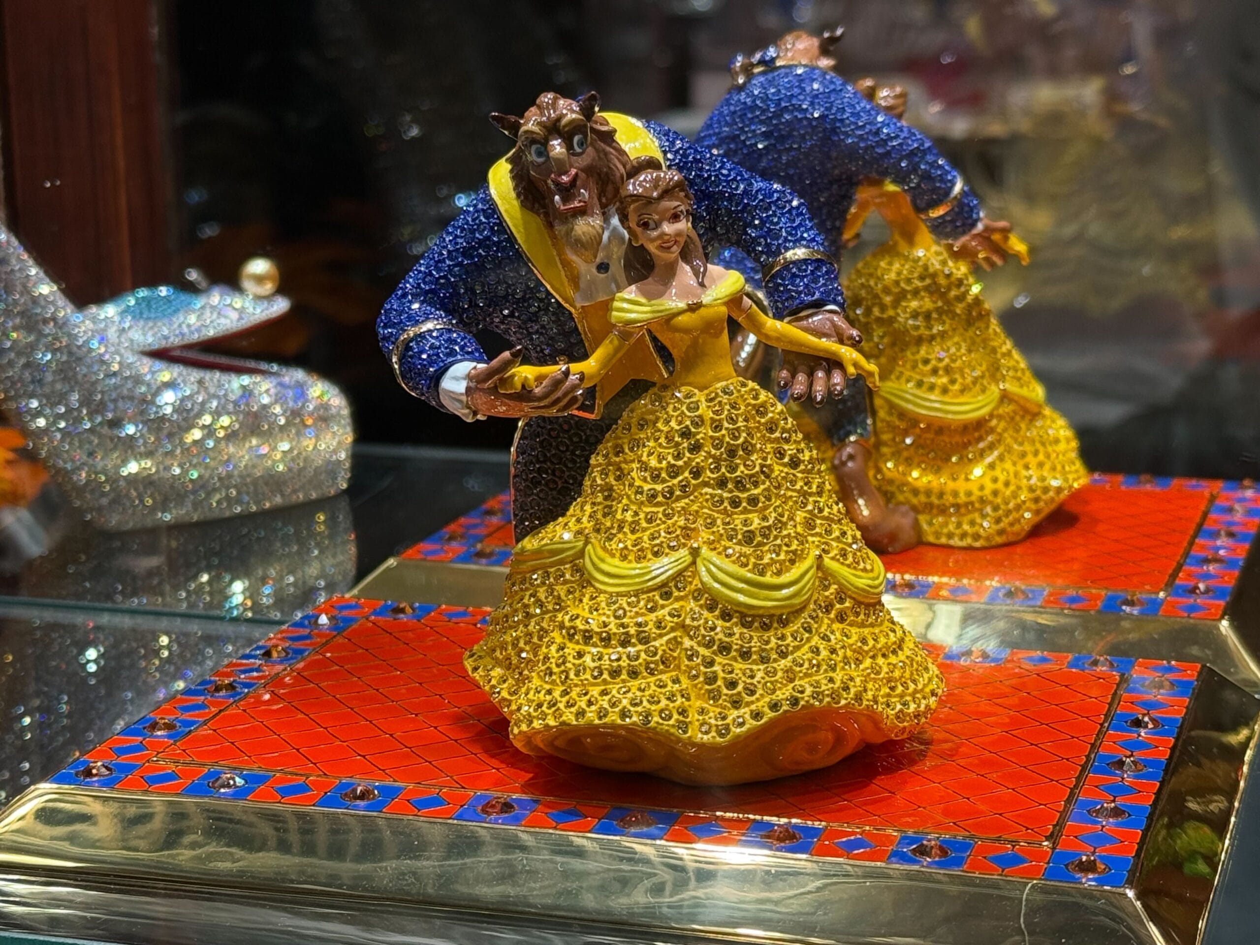 A jeweled Beauty and the Beast figurine on a red-blue tiled base, beside a glittery glass slipper, reminiscent of Disney Park displays.