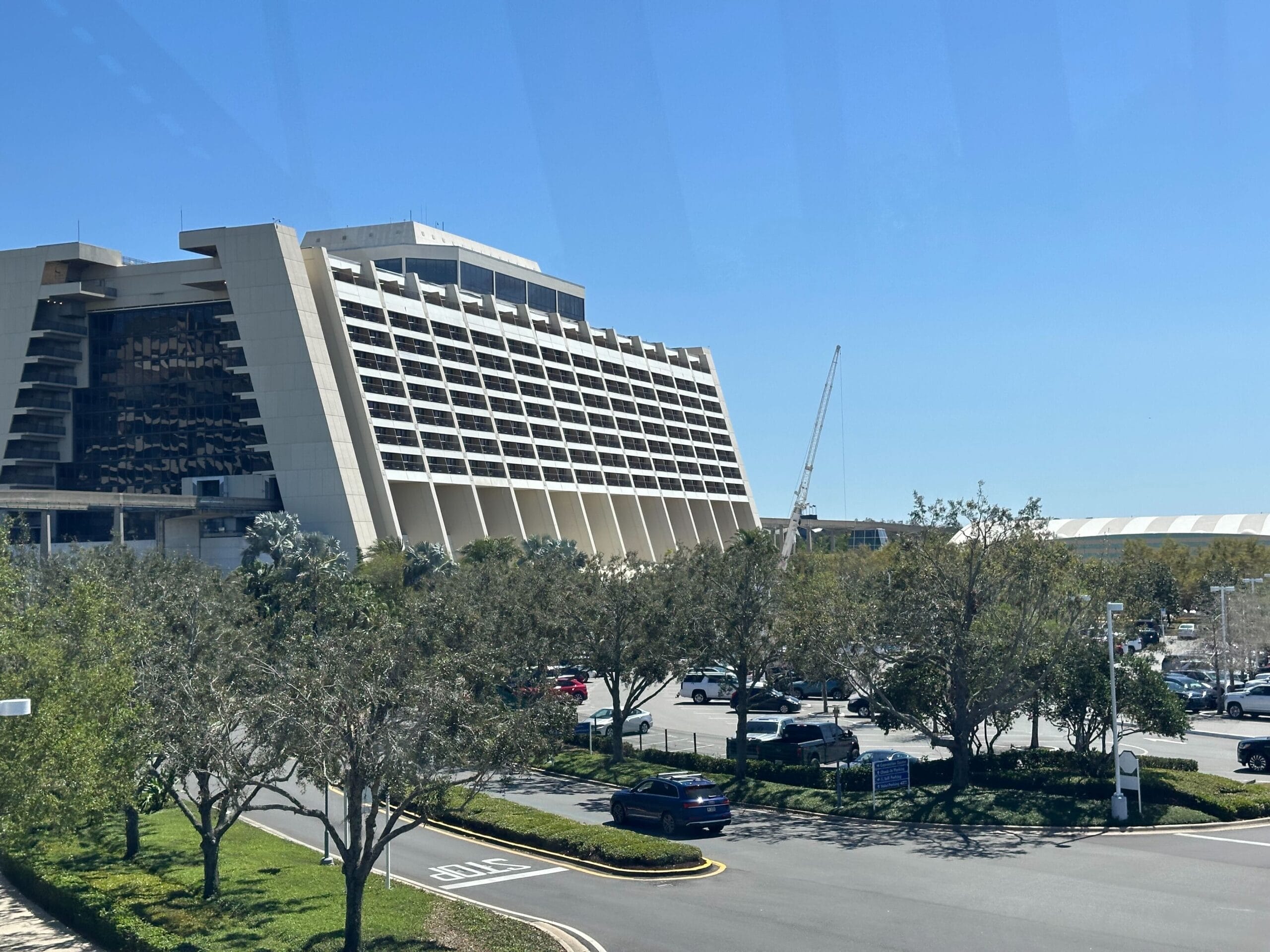 Disney's Contemporary Resort, sleek and modern, rises behind trees, parked cars, and a crane beneath a clear blue sky.
