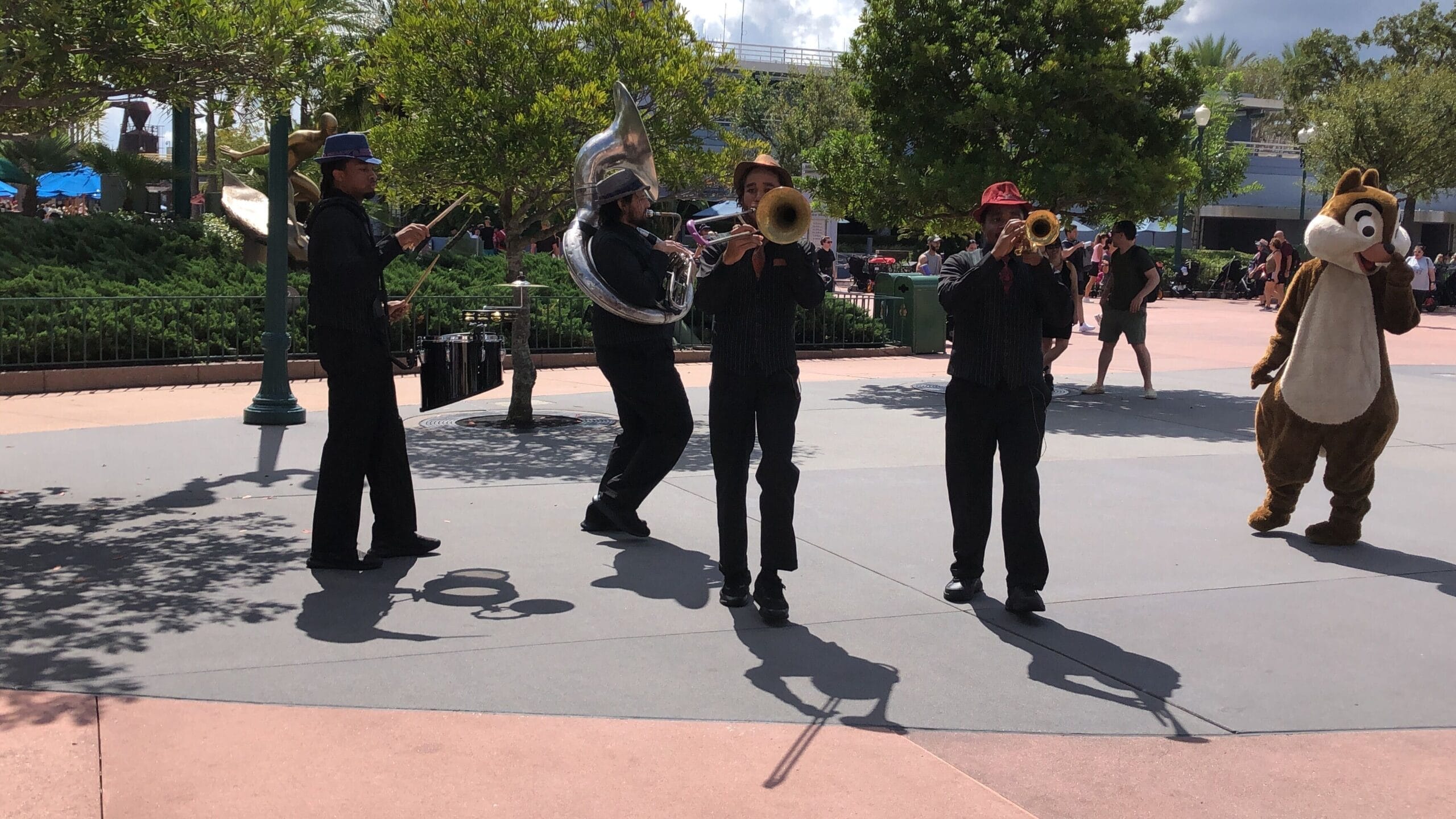 The HollyGroove Swinging Band plays brass outdoors at Hollywood Studios, with a Chipmunk mascot and crowds in the background.