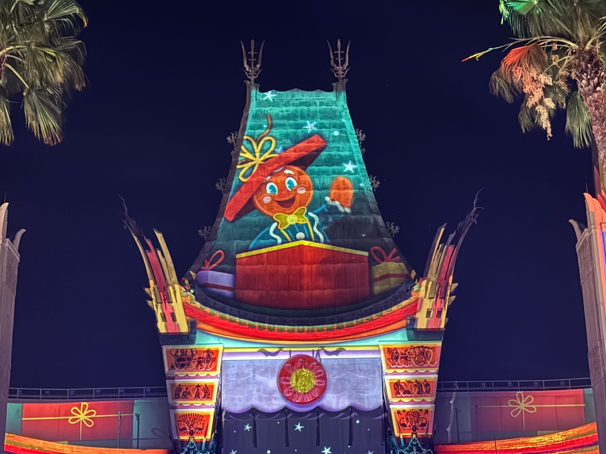 A cheerful cartoon gingerbread man is projected onto a vibrant building at Jollywood Nights 2025, framed by Disney palm trees.