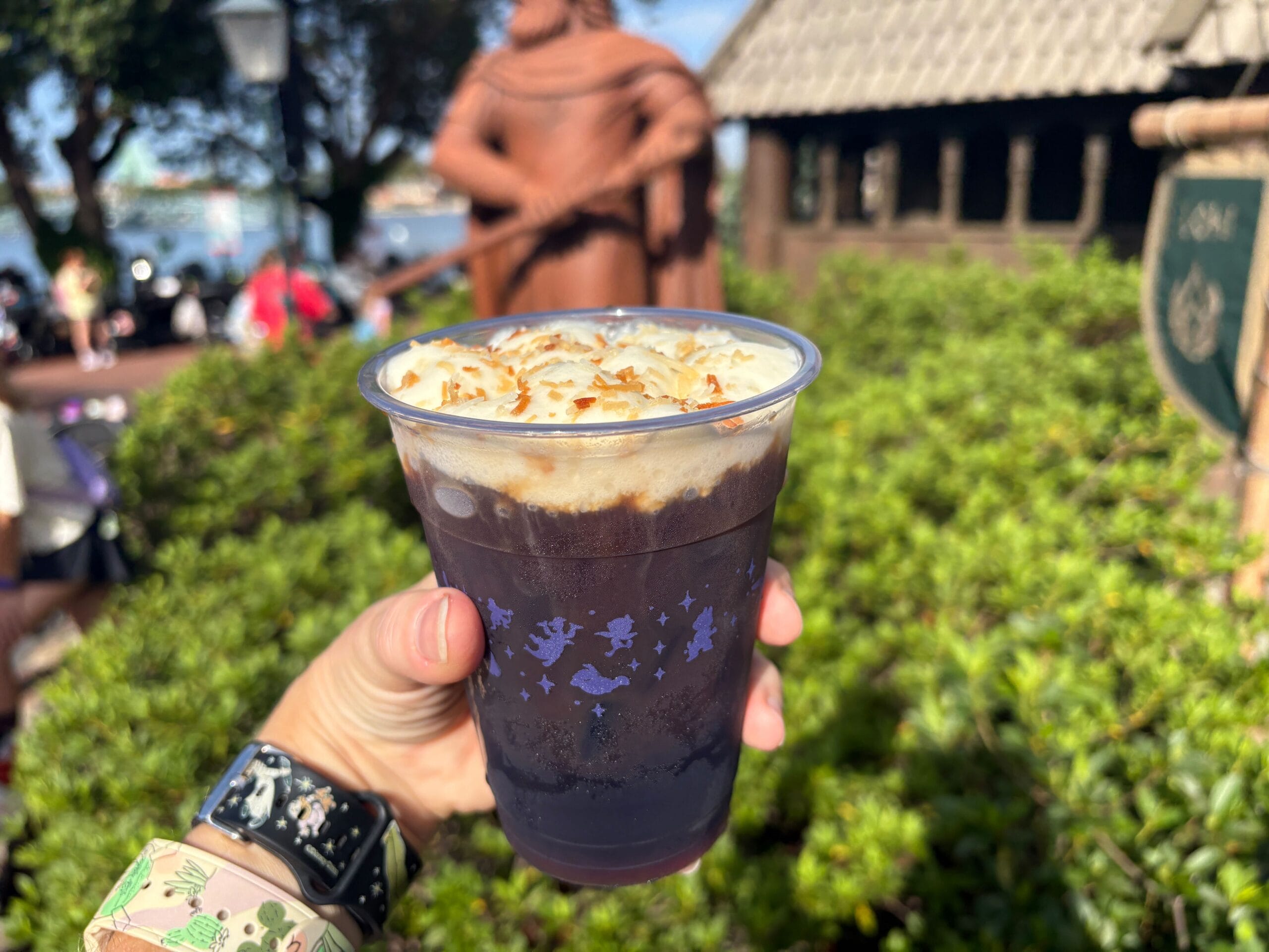 A hand holds a Cold Crumb Coffee with whipped cream and toasted coconut, inspired by EPCOT’s Norway Pavilion School Bread.