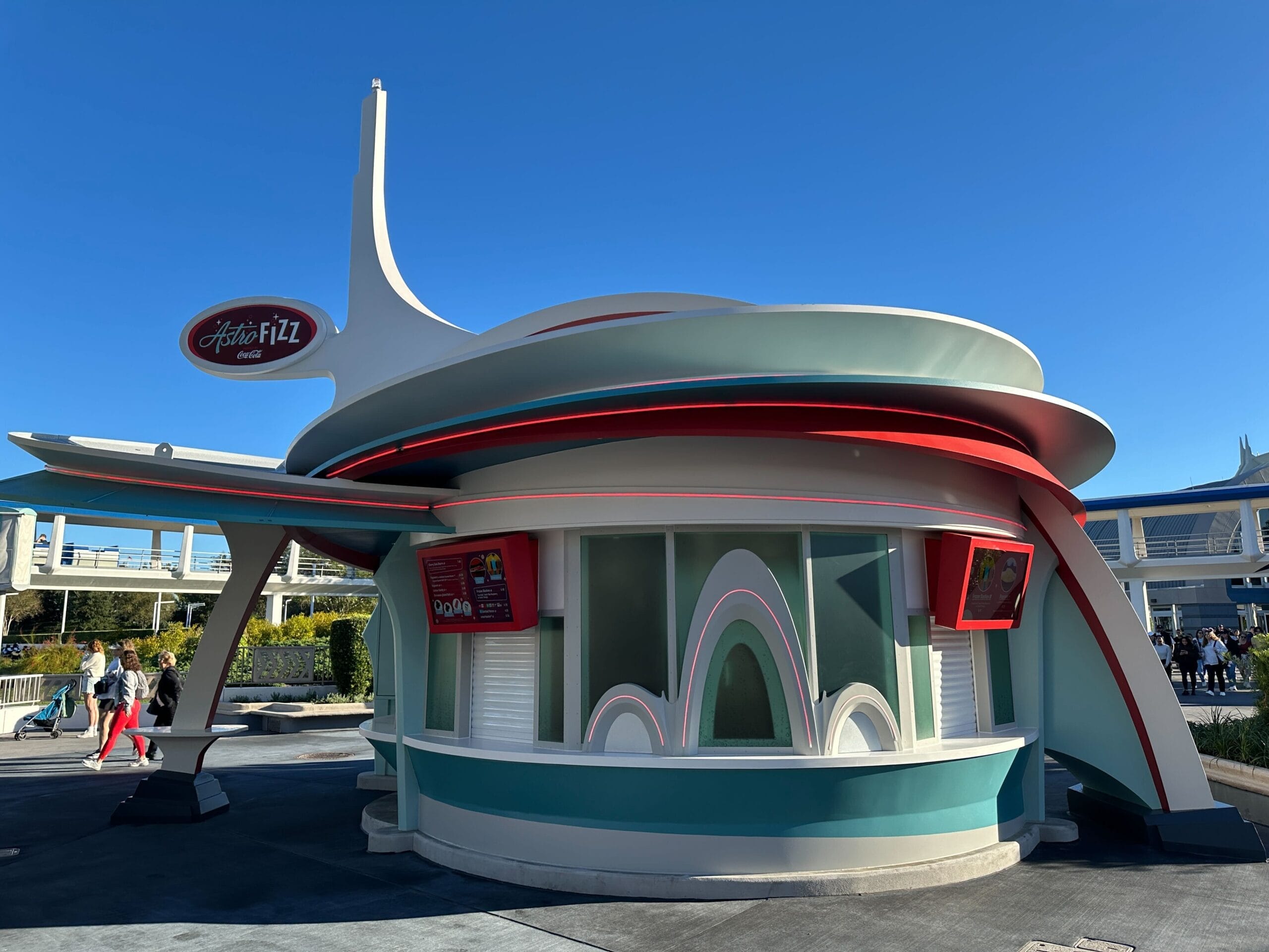 Astro Fizz snack stand with retro-futuristic curves and teal, white, red highlights—classic Tomorrowland vibes under blue skies.