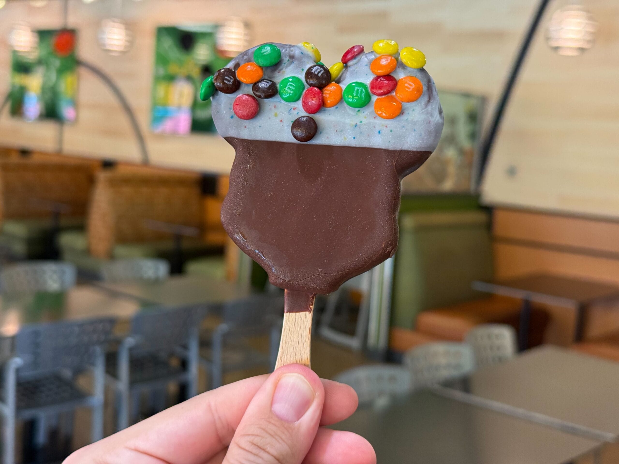 A hand holds a chocolate-dipped ice cream bar topped with colorful mini M&Ms in a modern café at a Disney park.