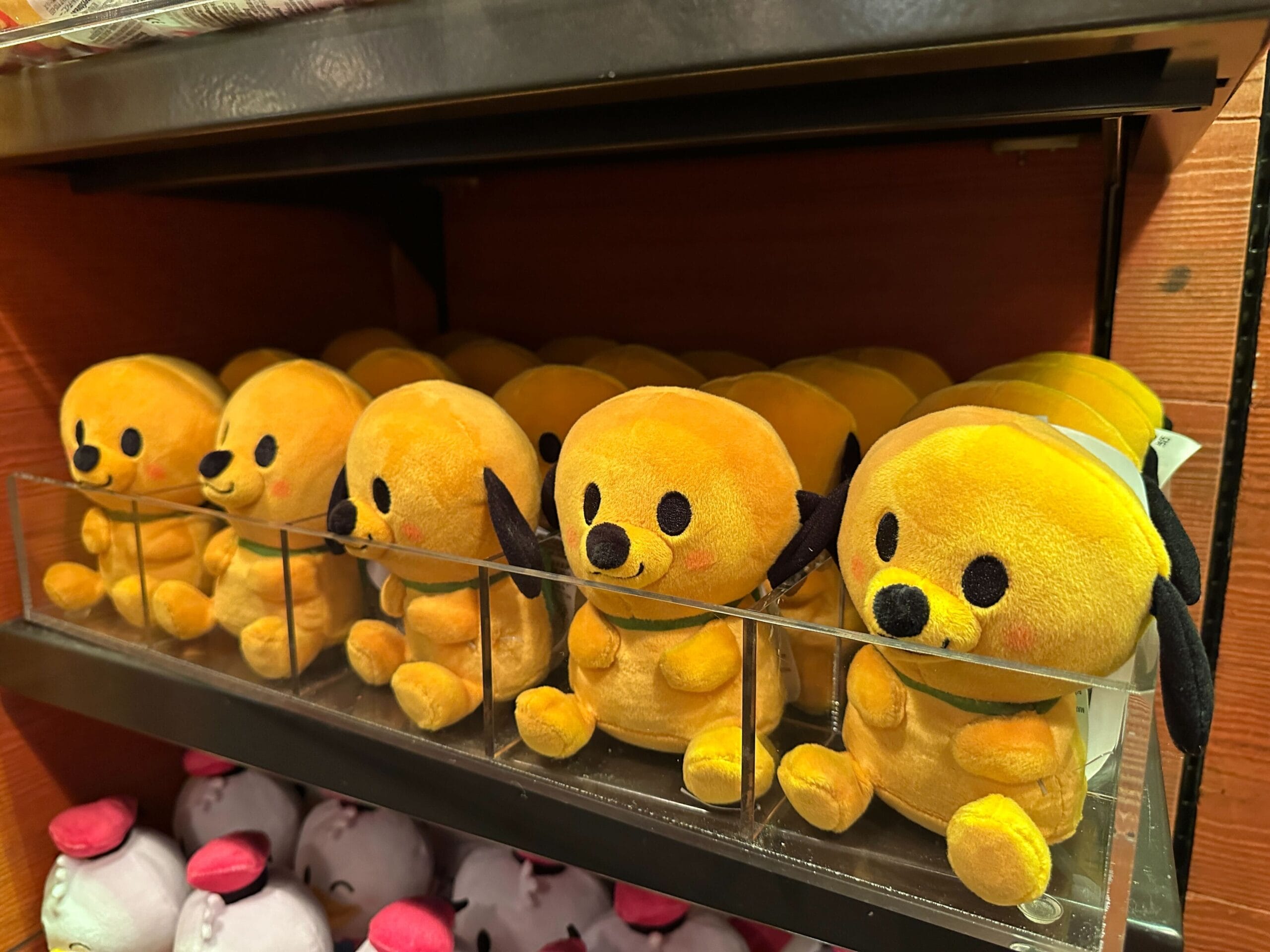Rows of yellow Pluto plush toys with black ears and rosy cheeks line a store shelf at a Disney park, ready for fans to purchase.