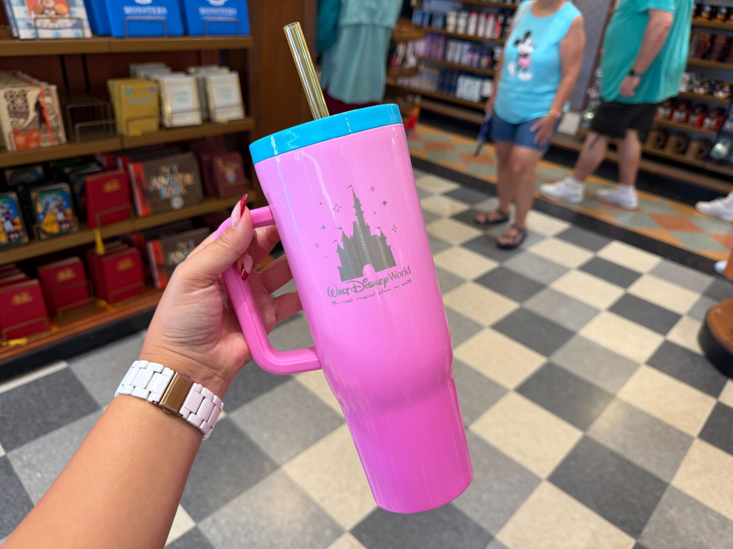 A hand holds a pink Walt Disney World tumbler with a straw in a gift shop full of Disney souvenirs and books, no castle visible.