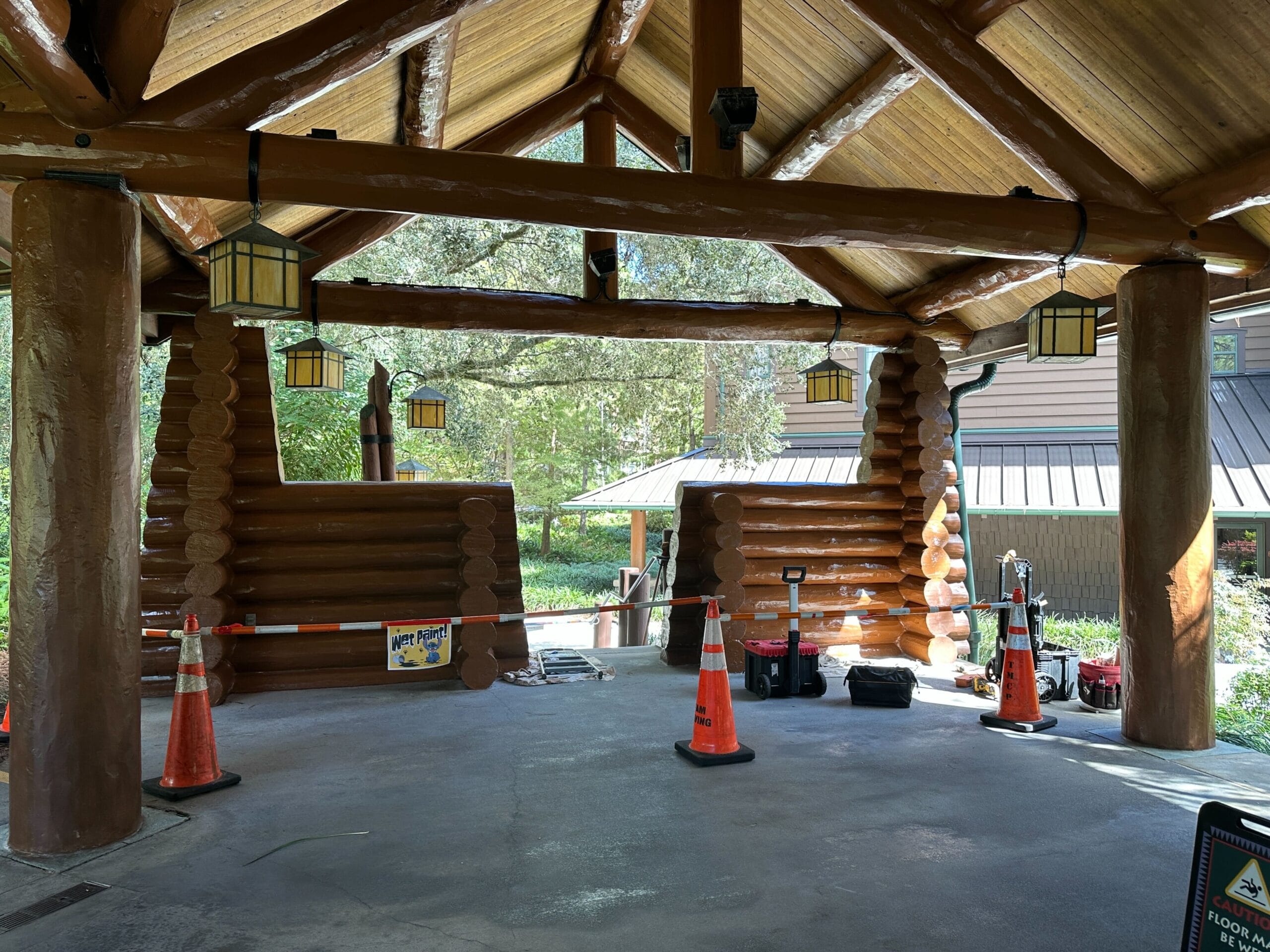 Rustic log cabin pavilion with orange cones, tools, and lanterns; lush greenery evokes Frontierland ambiance at Disney parks.