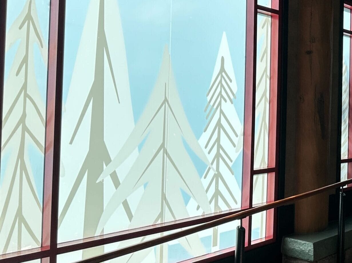 Guests stroll along a wooden hallway with frosted tree designs on the windows, sunlight streaming in like at Disney’s Wilderness Lodge.