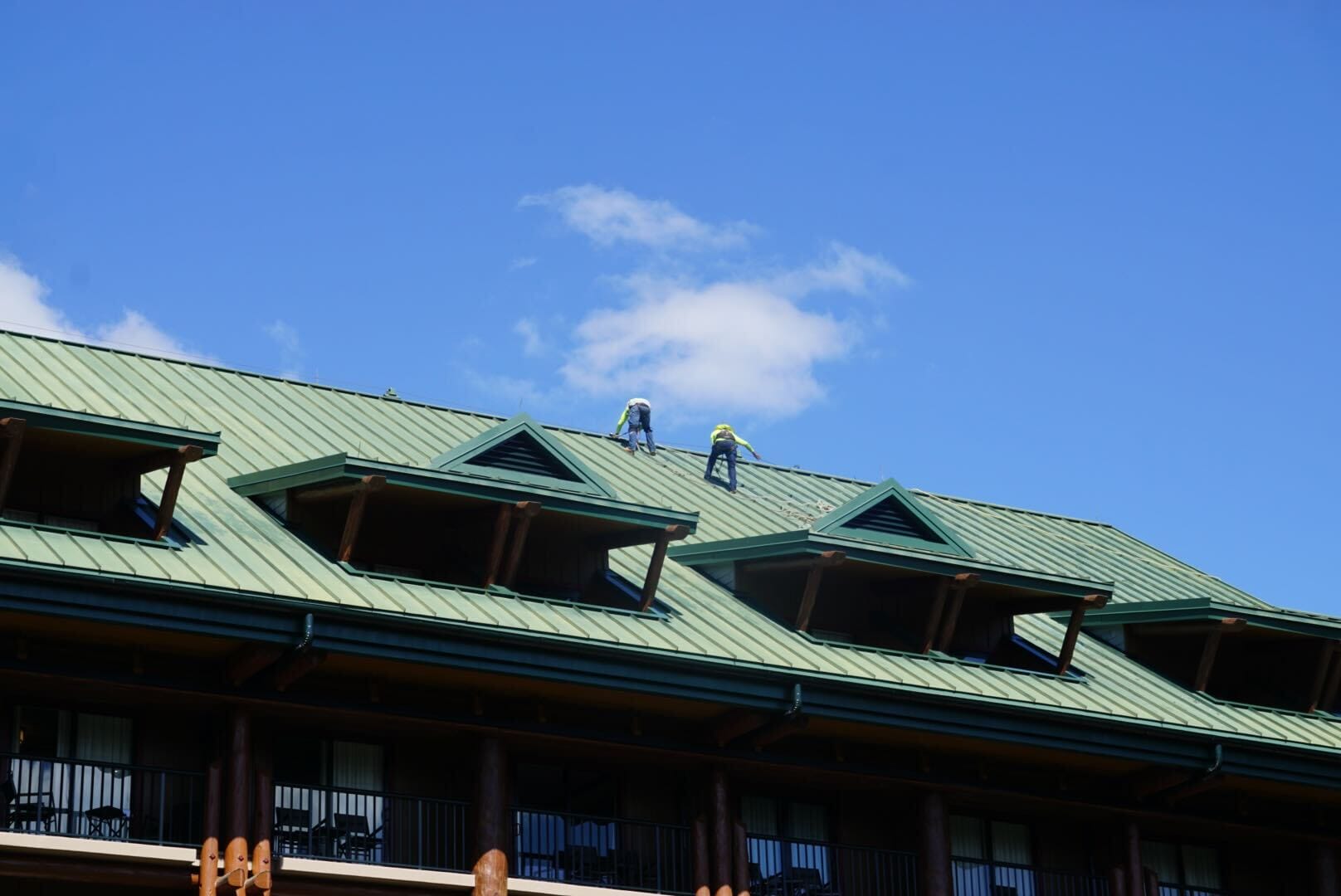 Two workers in safety gear work atop a green metal roof under a blue sky, reminiscent of rooftops seen in Disneyland parks.
