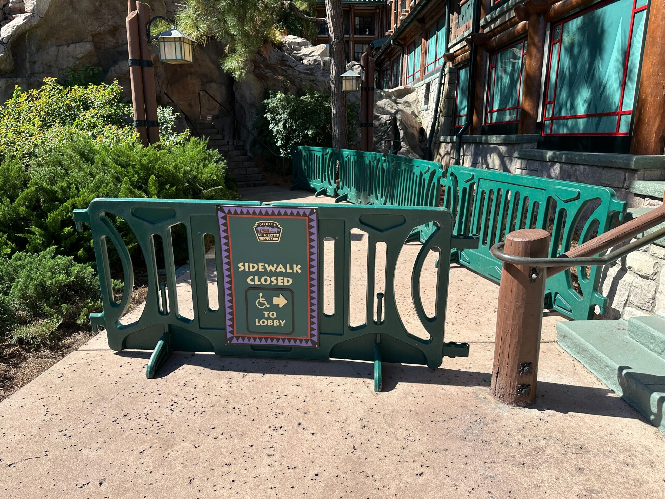 Green construction barriers block the sidewalk, with a sign saying "Sidewalk Closed, to Lobby" and a left arrow, Disney park style.
