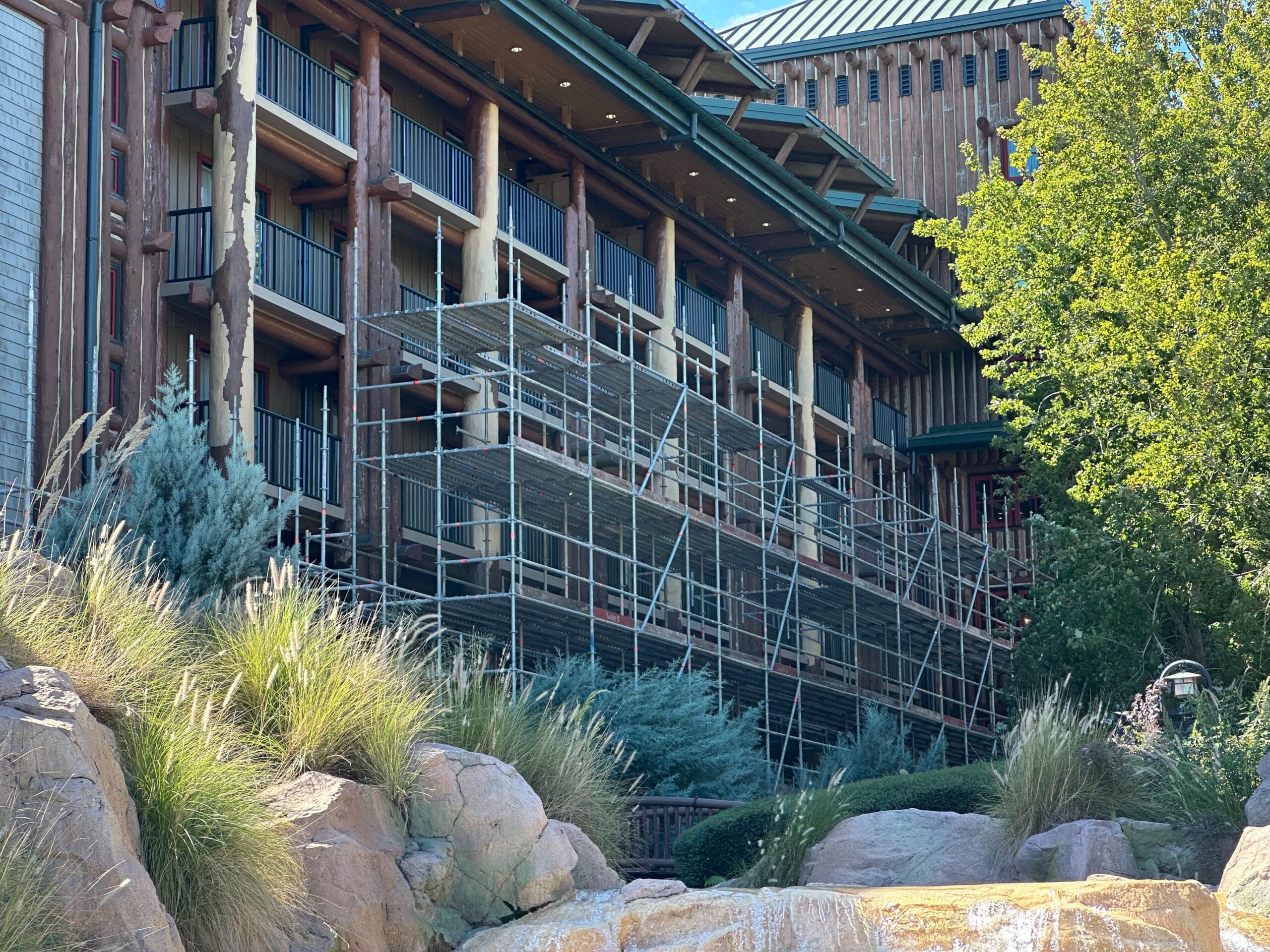 Scaffolding covers multiple floors of a rustic hotel, nestled among lush greenery and large rocks, reminiscent of Disney's Wilderness Lodge.