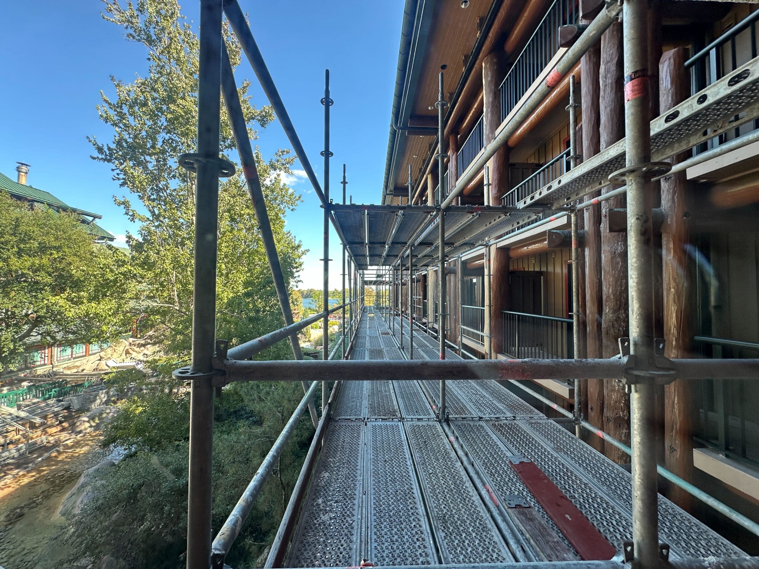 Scaffolding lines the side of a building, possibly during a refurbishment like those seen on Disney park castles, with trees and blue sky behind.