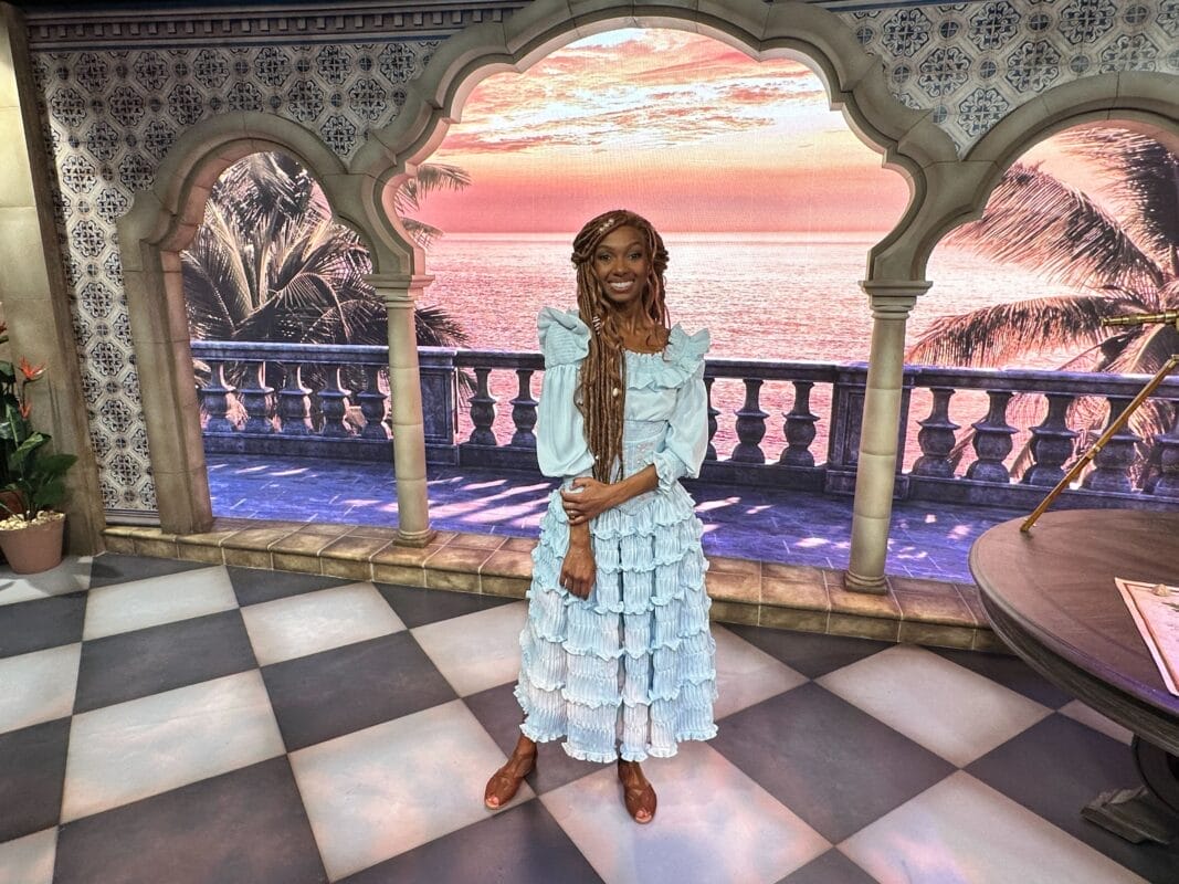 A woman in a ruffled light blue dress stands on tiles at Hollywood Studios Ariel Meet & Greet, sunset and pillars behind her.