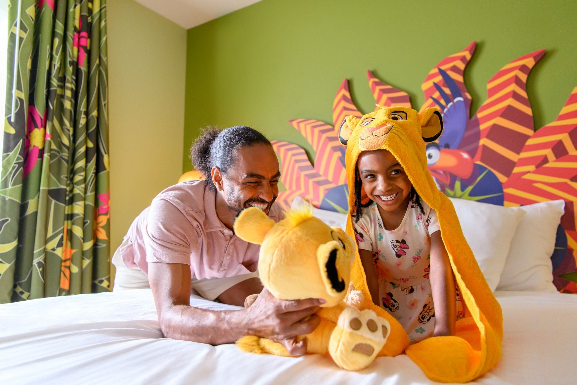 A man and a girl in animal hats smile and play with a plush toy, enjoying cool kid summer vibes in a colorful room.