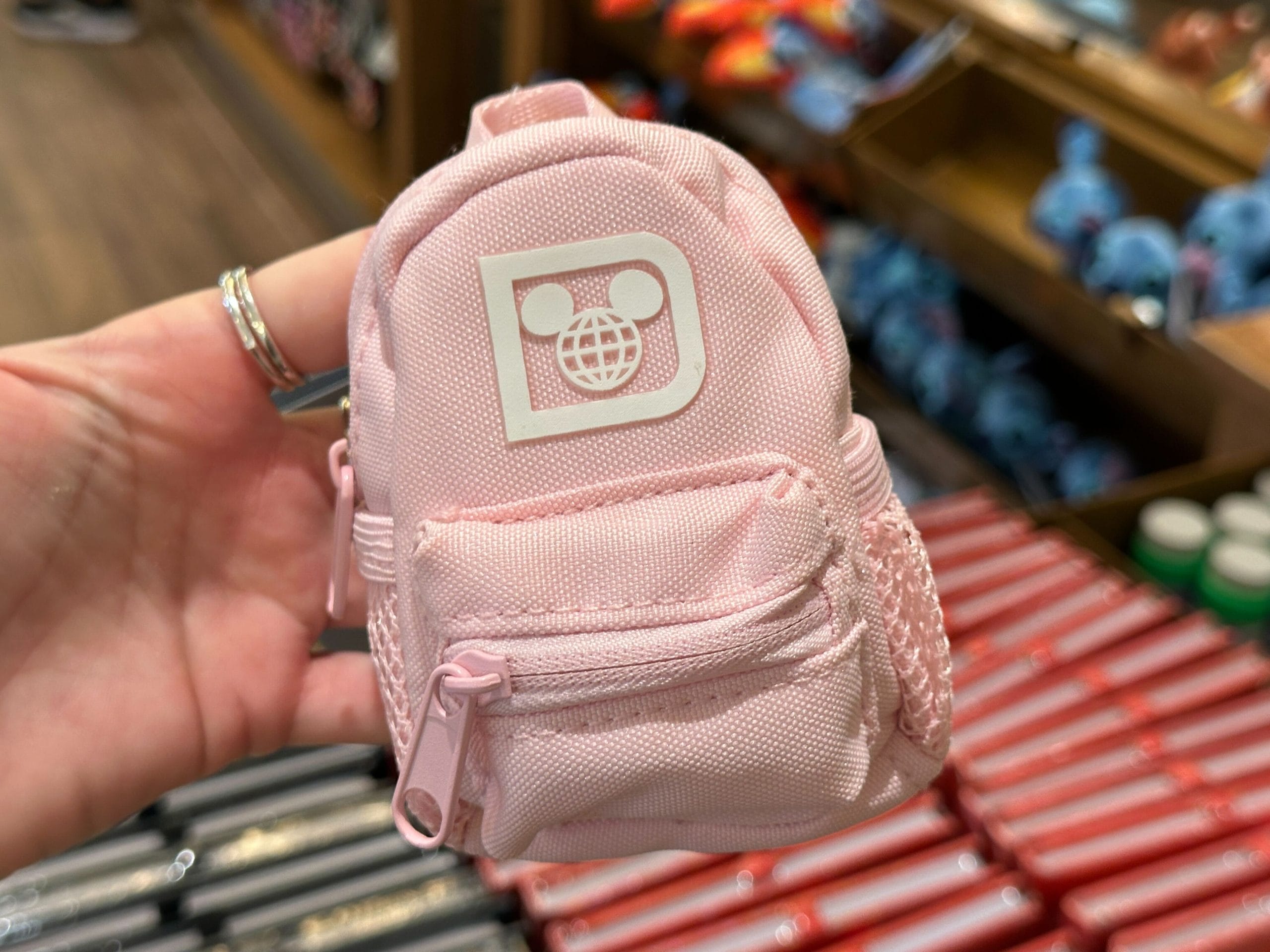 A hand holds a small pink backpack keychain charm with a white Walt Disney World "D" logo.