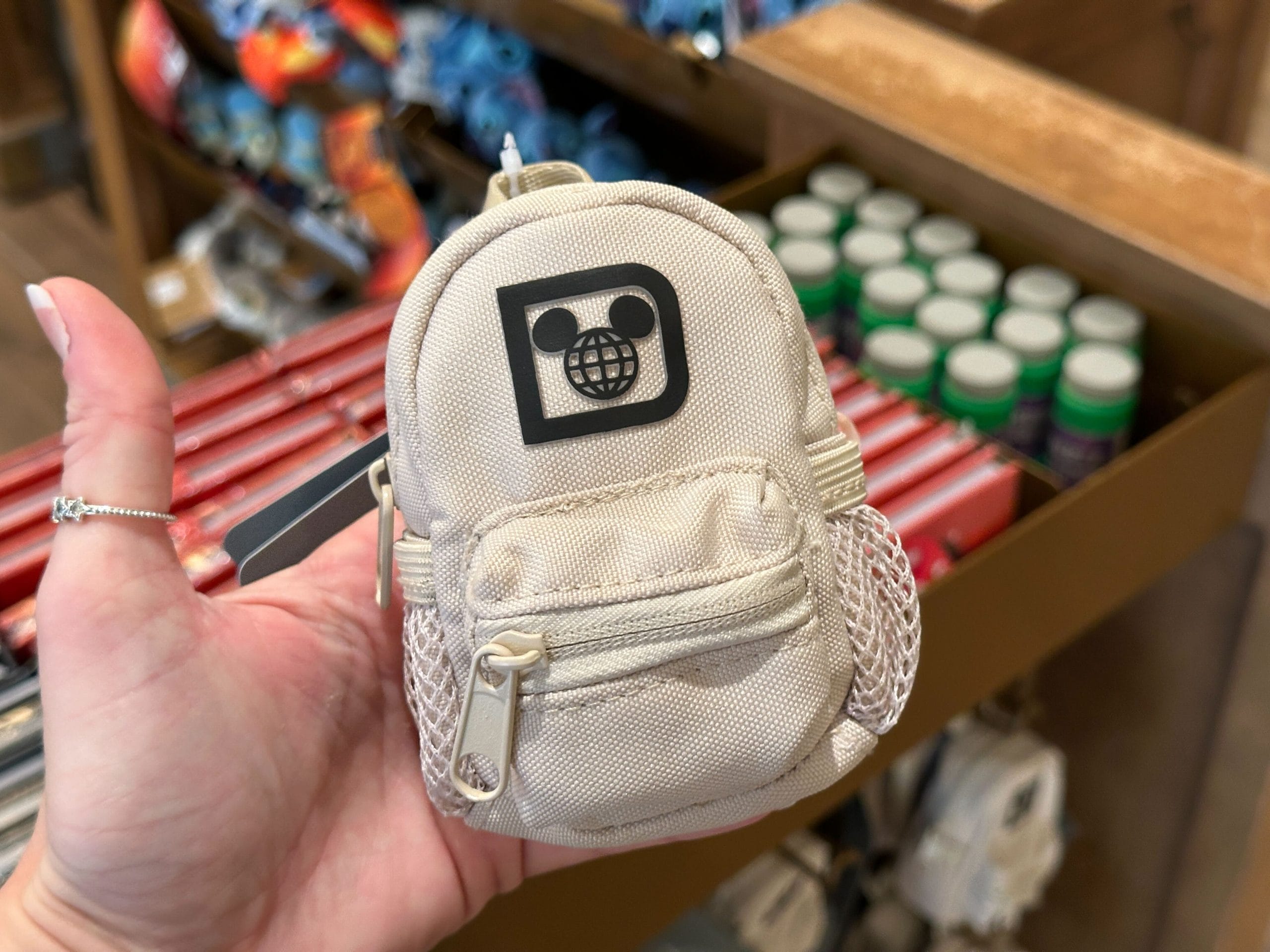 A hand holds a small white backpack keychain charm with a black Walt Disney World "D" logo.