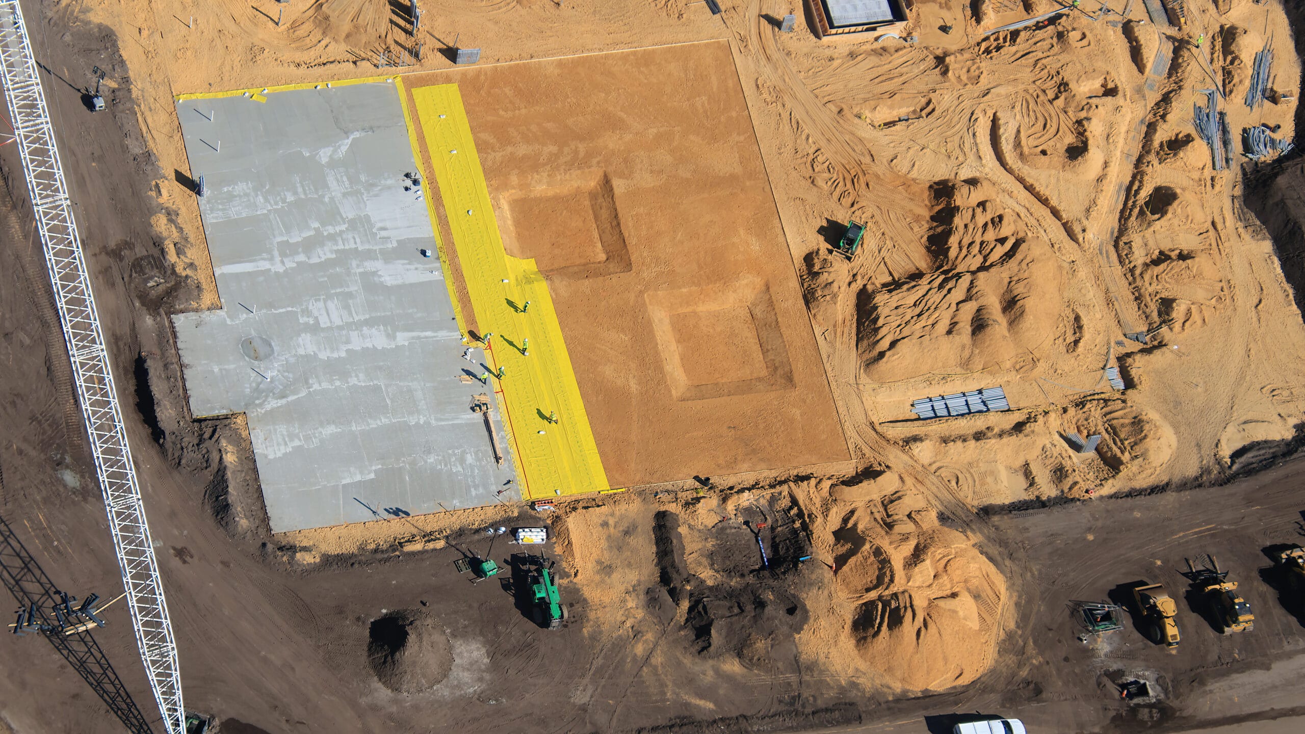 At the site of the coaster, we see that a concrete floor section has been poured on the left with a moisture barrier beside it. Electrical conduits are being buried, as evidenced on the right side. We can also see that some concrete flooring has already been formed and placed. You can see these where there are perfect squares in the sand in the center of the photo.