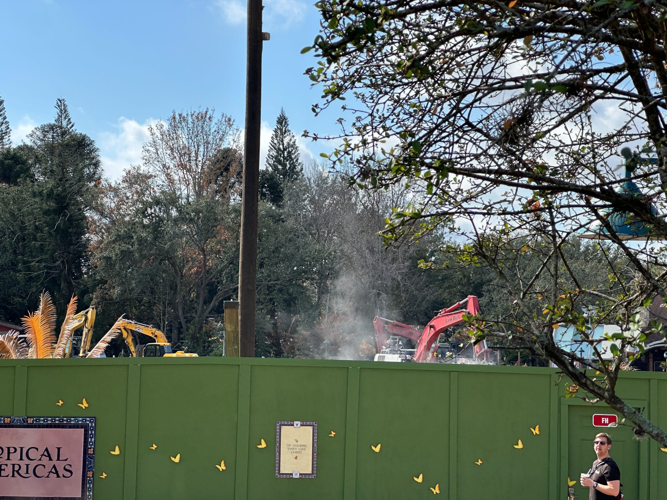 Over the construction wall, we see several excavators, rocks, and upturned dirt. There are stacks of wood and some concrete blocks likely to be installed or moved in the coming days. Dust floating up above the wall indicates active excavation being done by crews.