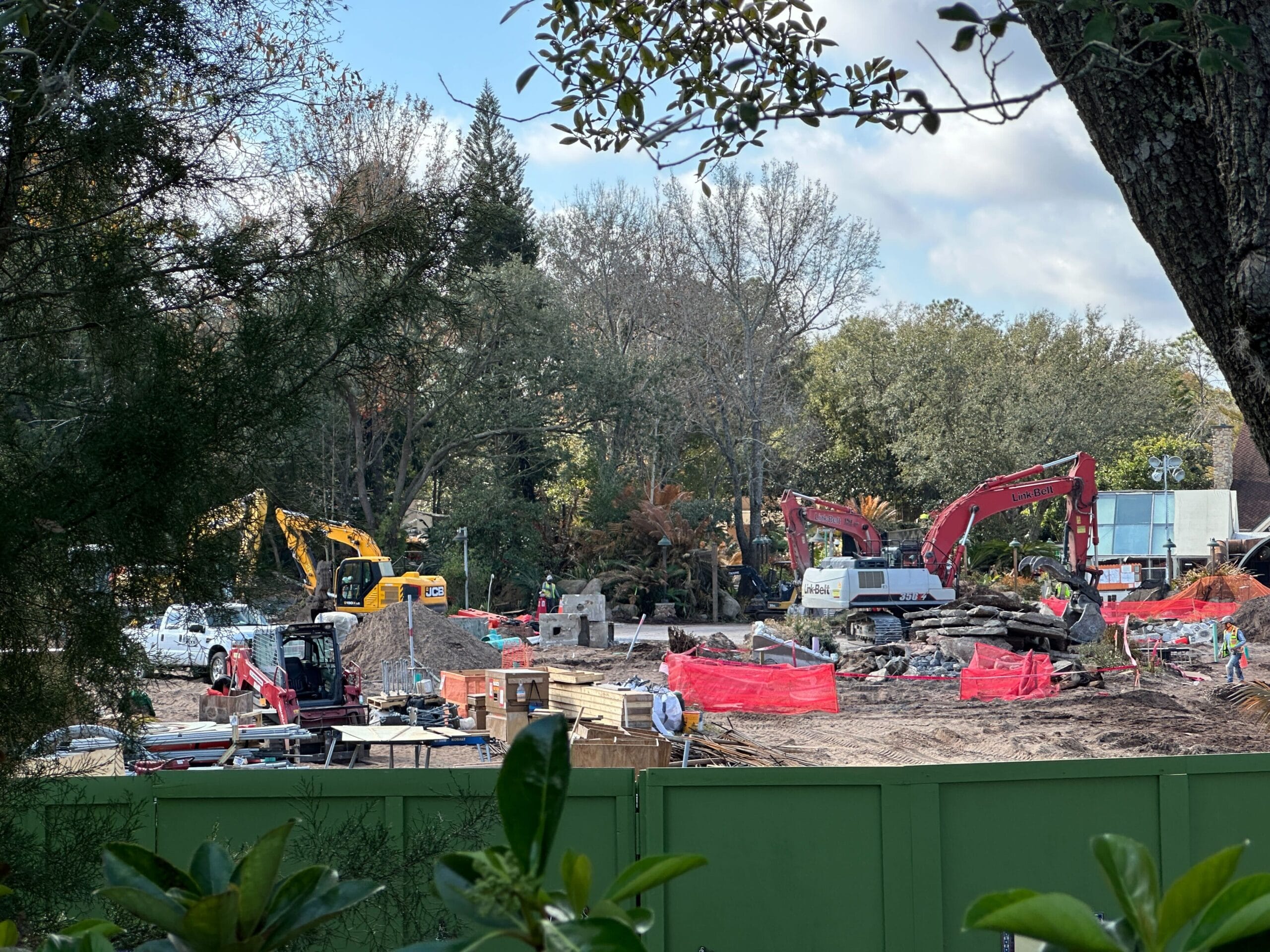 Over the construction wall, we see several excavators, rocks, and upturned dirt. There are stacks of wood and some concrete blocks likely to be installed or moved in the coming days. Dust floating up above the wall indicates active excavation being done by crews.