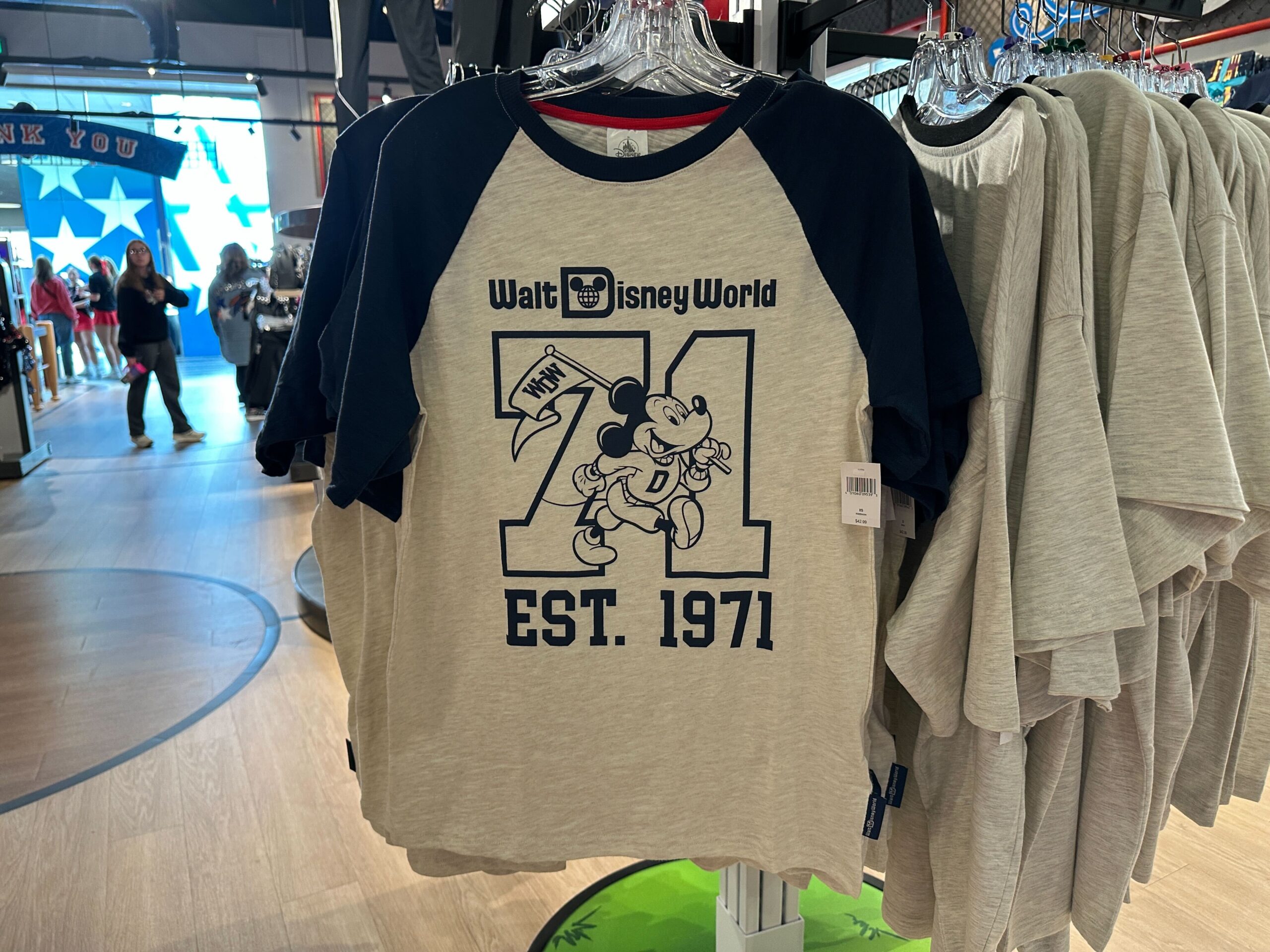 Raglan tee with artwork on the chest featuring Mickey holding a "WDW" flag and wearing a "D" sweater. He's marching across a large "71." The retro Walt Disney World wordmark and "EST. 1971" frame the design.