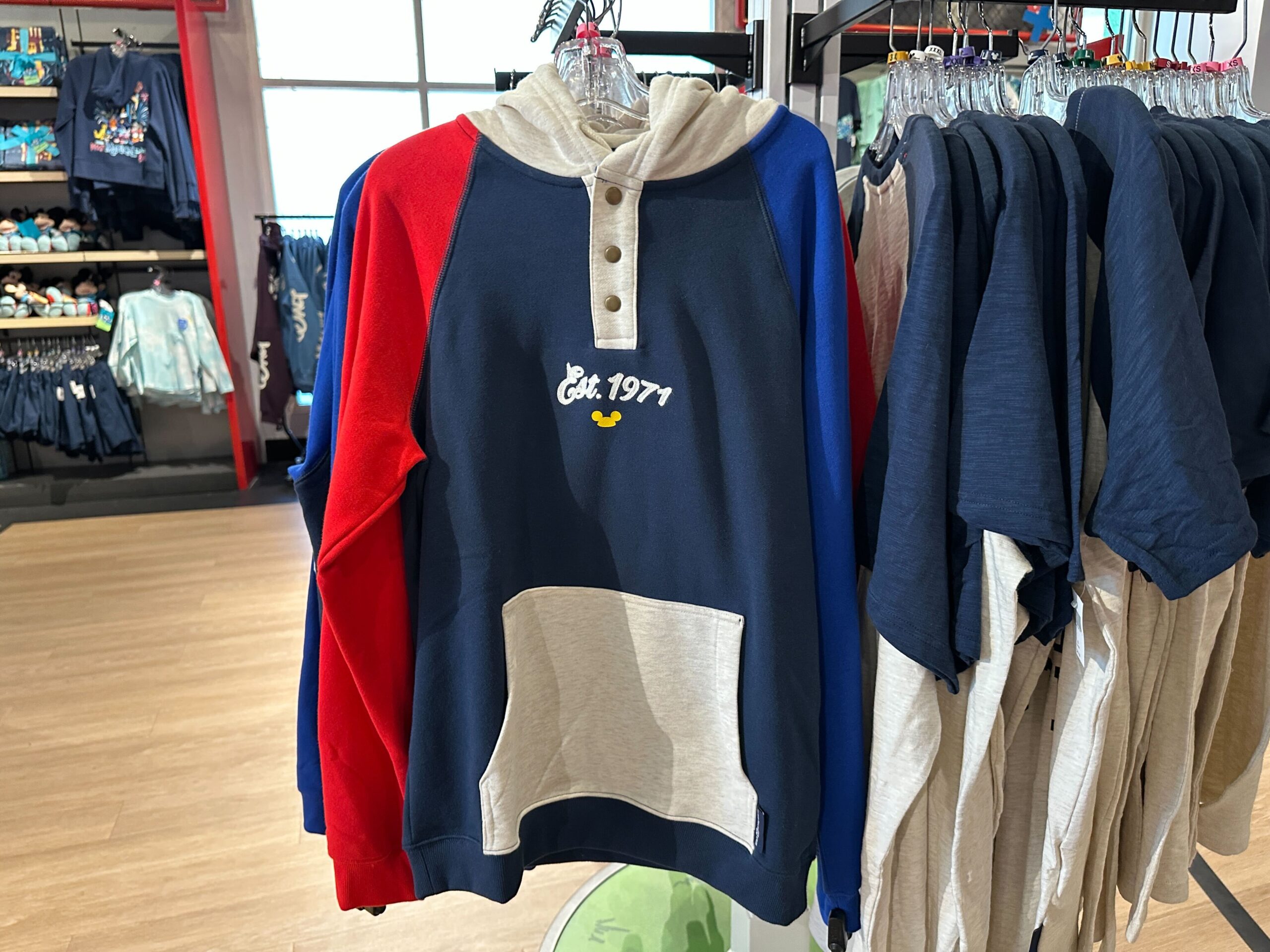 The hoodie is color-blocked with a dark blue torso, bright blue left sleeve, and bright red right sleeve. The hood and kangaroo pocket are heather gray. A trio of buttons leads down the shirt.