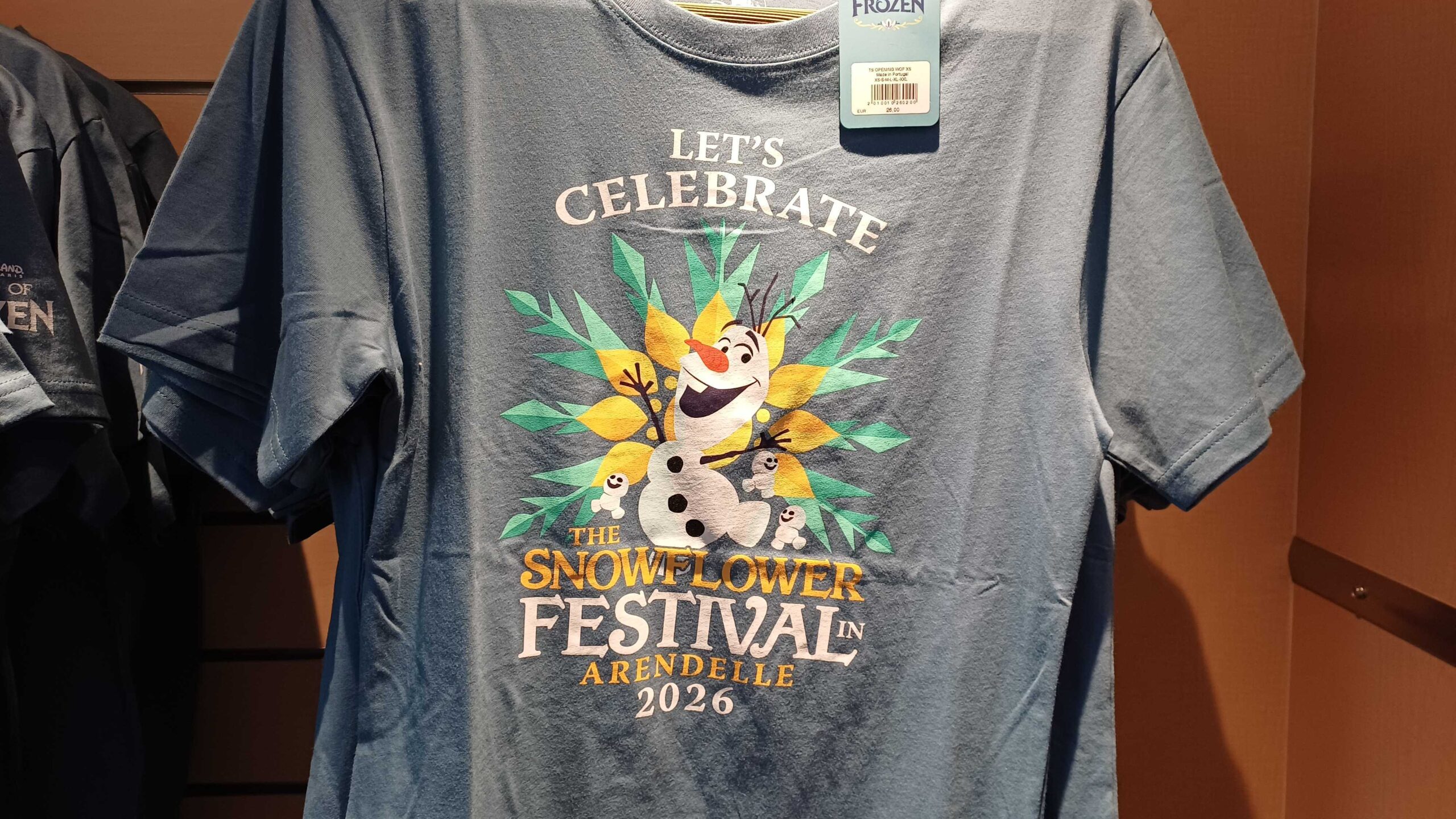 Similarly, Olaf and the Snowgies are front and center again for this Snowflower Festival shirt. They are dancing in front of a green and yellow snowflake. Above them are the words "Let's celebrate" and "The Snowflower Festival in Arendelle 2026" are beneath. The back is plain.
