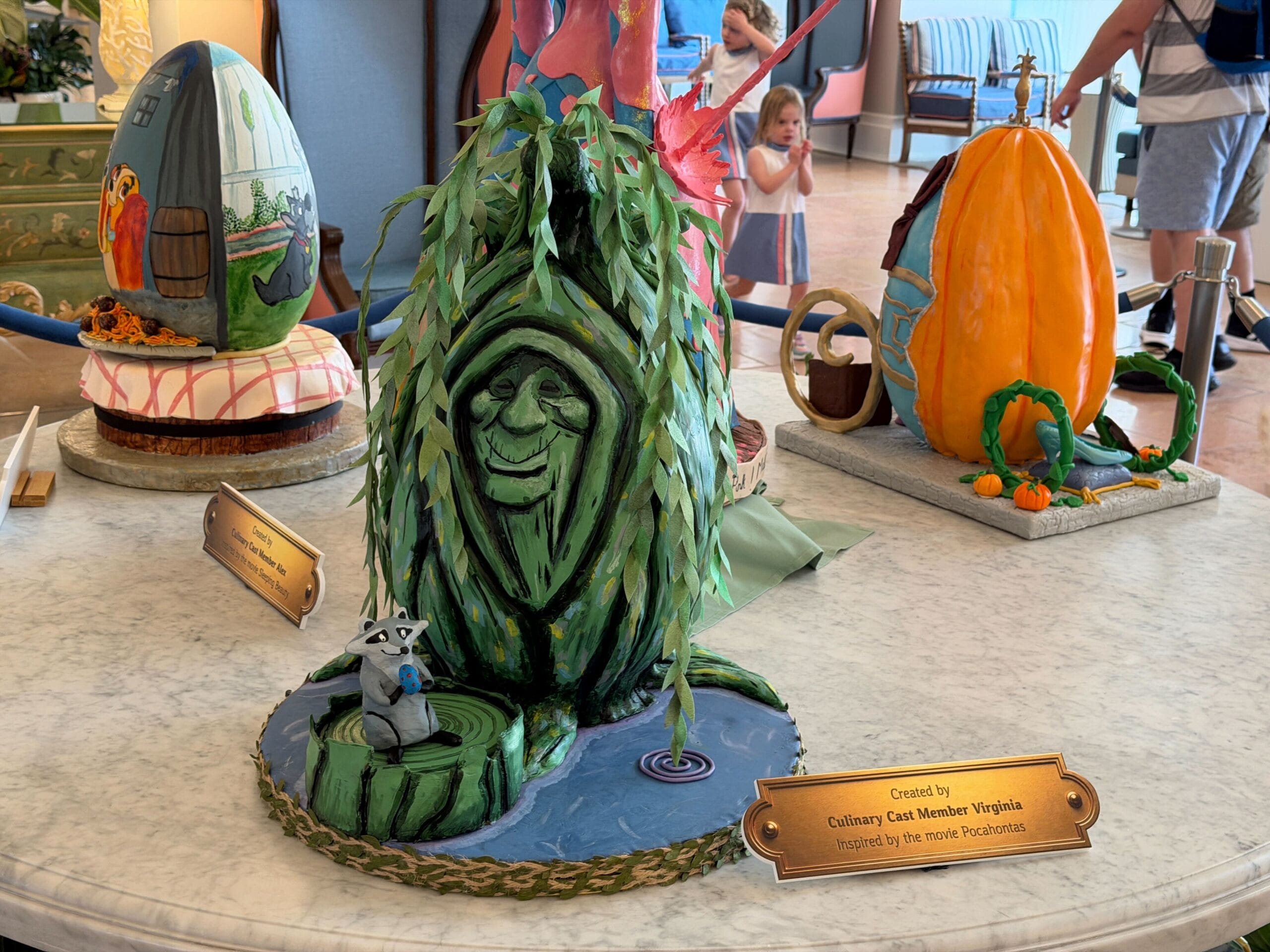 The second egg is inspired by Pocahontas, featuring Grandmother Willow in the shape of an egg. Meeko the raccoon is holding a small egg on the base, standing on a log. Part of Grandmother Willow is touching the water, which is the base of the display.