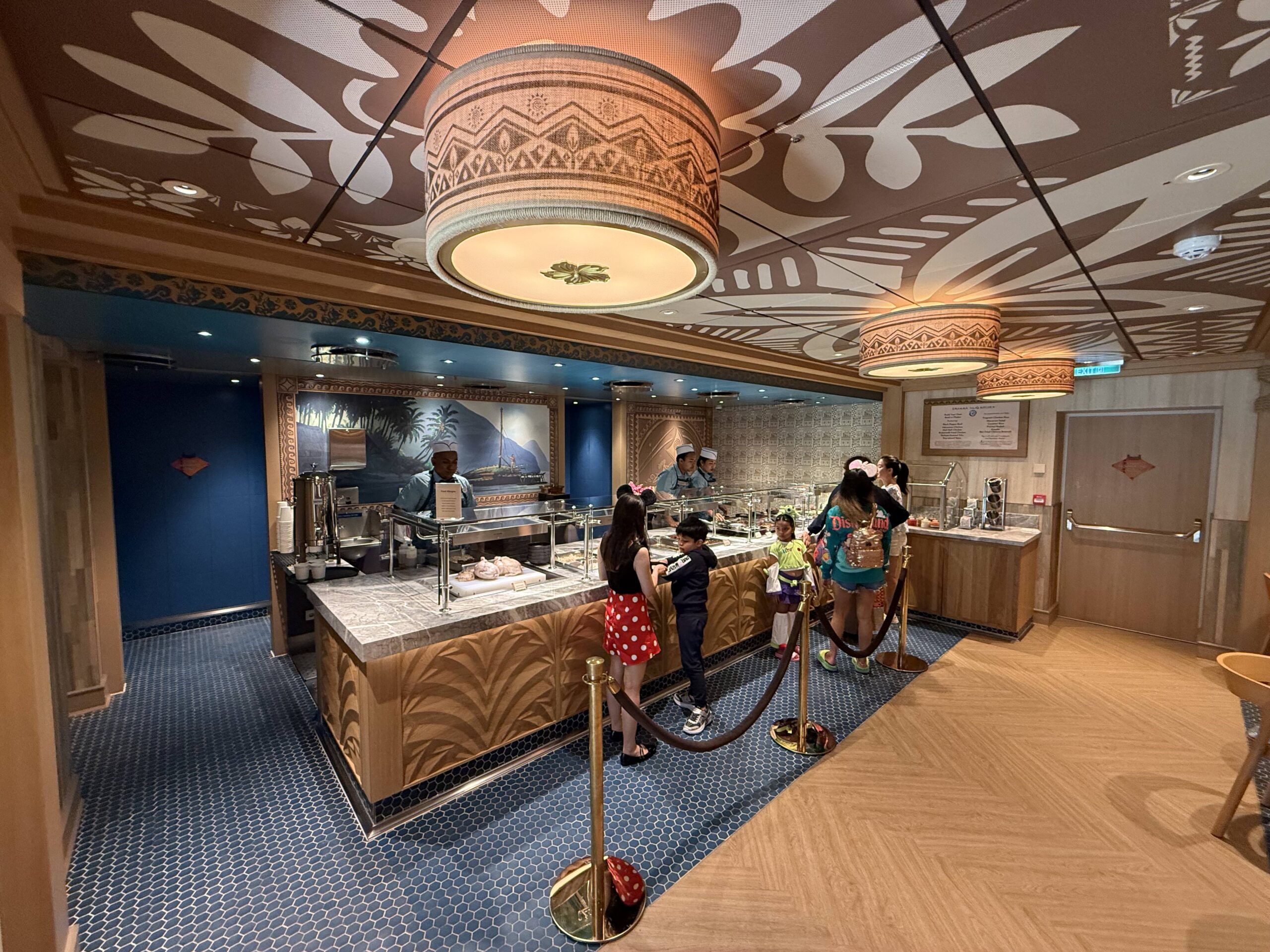 Inside, there is a small buffet area where crew members serve up the food to guests. Behind the food area is a large mural featuring an ocean scene. Dark blue paneling and patterned wallpaper make up the walls.