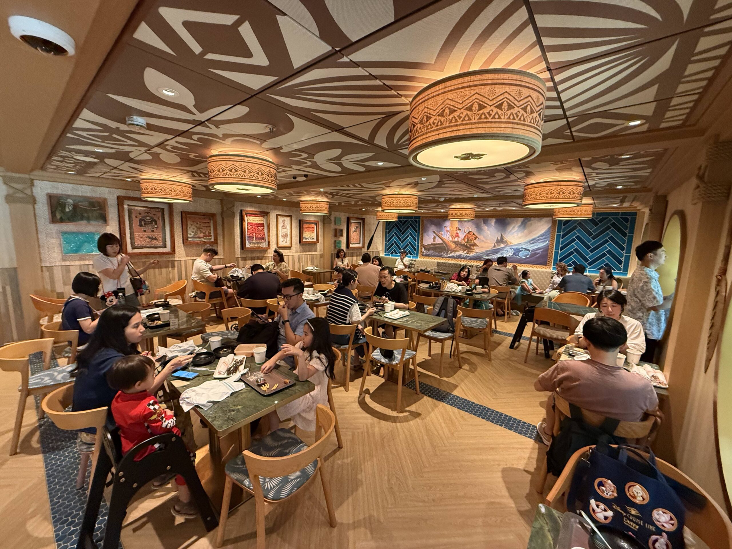 The seating area is a combination of about 20 two- and four- top tables. The flooring in here is mainly the herringbone vinyl plank, but there are strips of the blue tiles. In the back is a large mural scene of Maui and Moana on their boat in the ocean. Blue and teal chevron tile plank the mural on either side. Along the other wall is a variety of portraits and nods to the movie.
