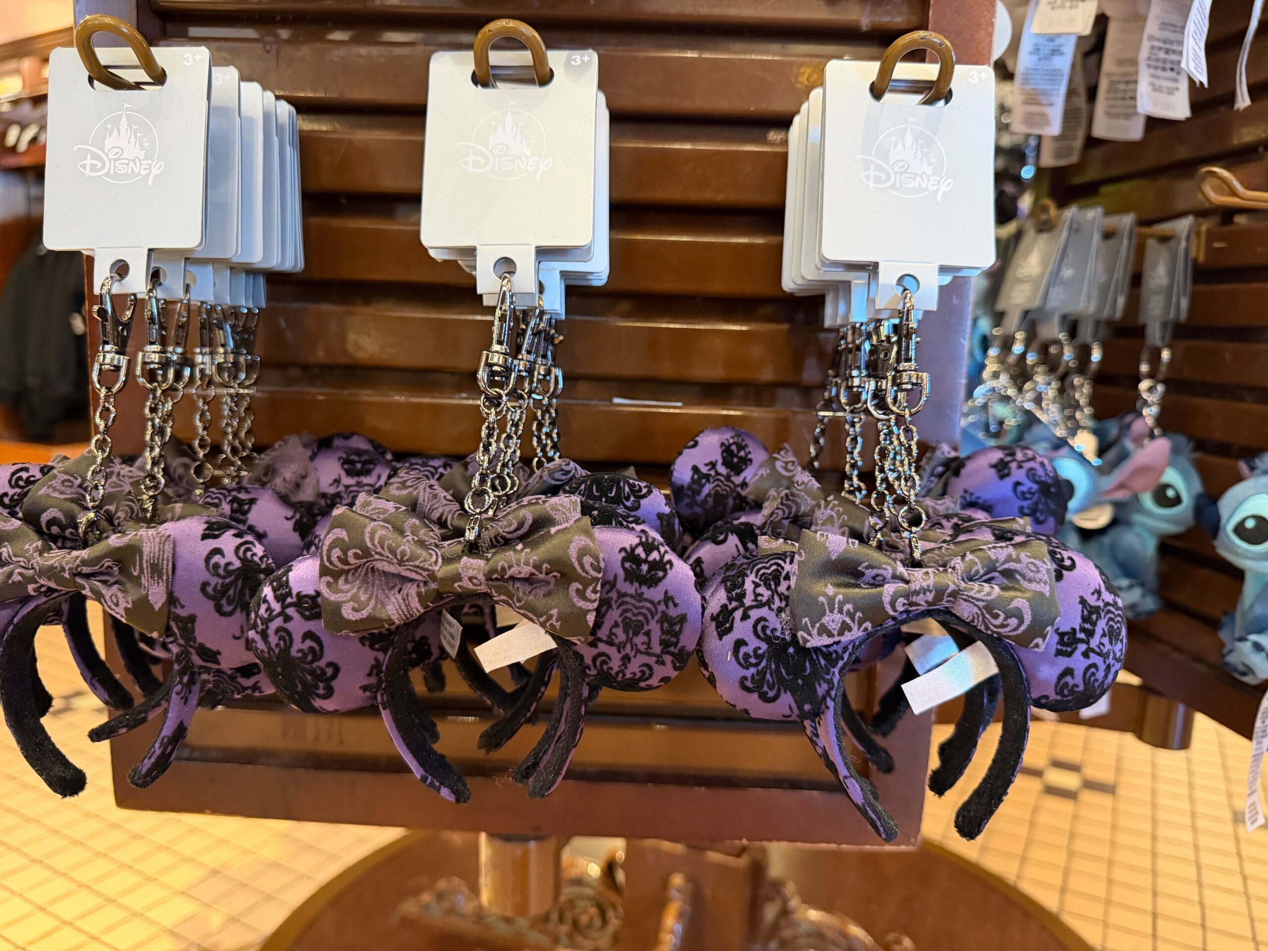 This tiny ear headband hangs from a silver-tone chain with a lobster claw clip. The ears are purple with a black velour pattern inspired by The Haunted Mansion's wallpaper. The bow is black with a purple design.