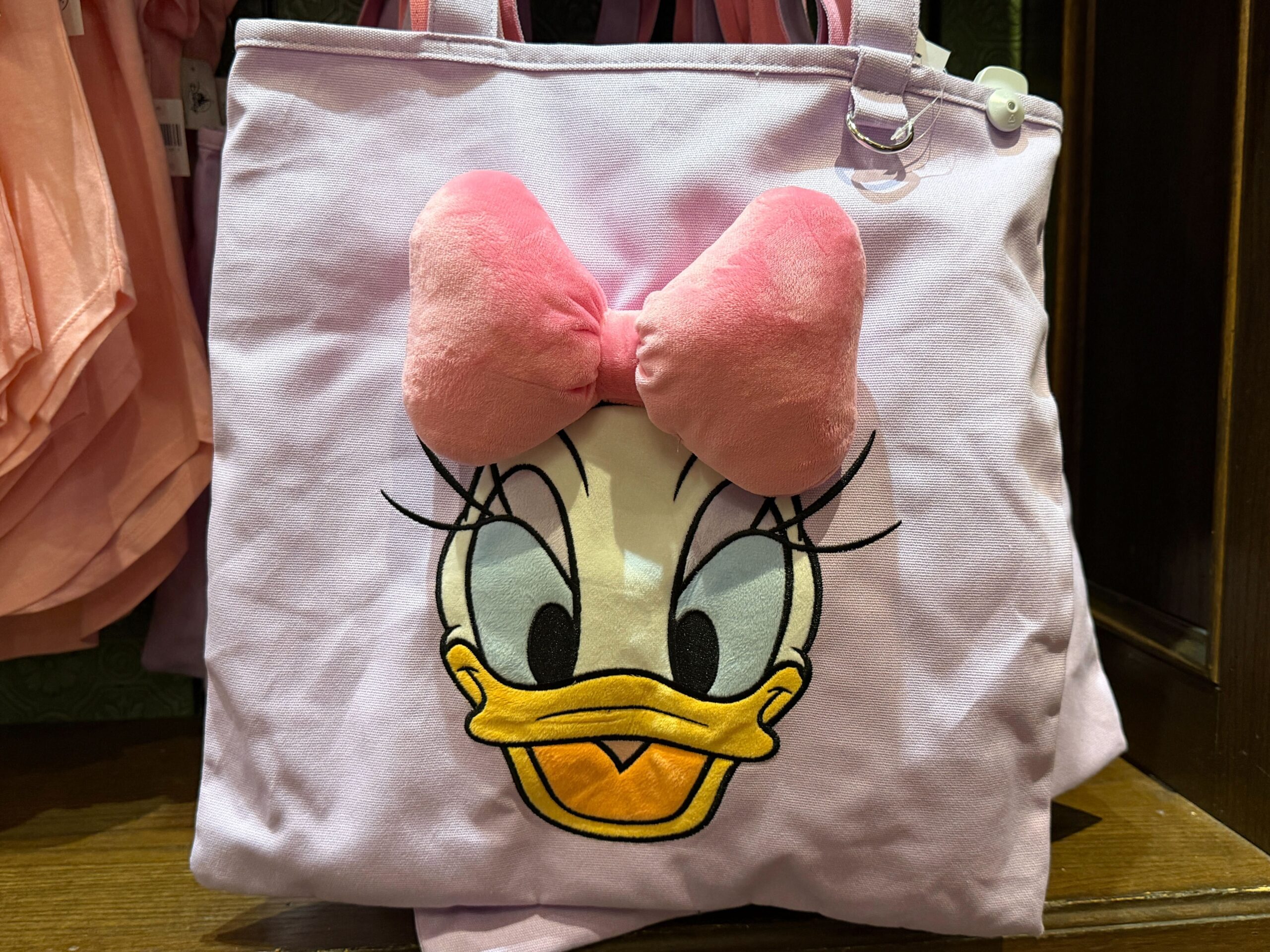 Daisy's face is embroidered on the front with a large pink plush bow. A metal ring below one strap is perfect for hanging your ear headband keychain.