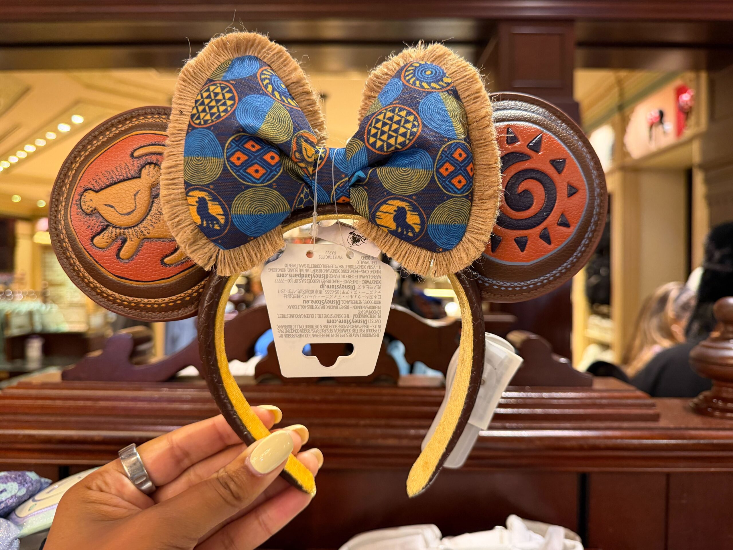 The padded ears on this accessory are made of brown debossed pleather. On the right ear is Rafiki's Simba drawing against an orange circle with a yellow border. The other ear features a black sun design with a blue border.