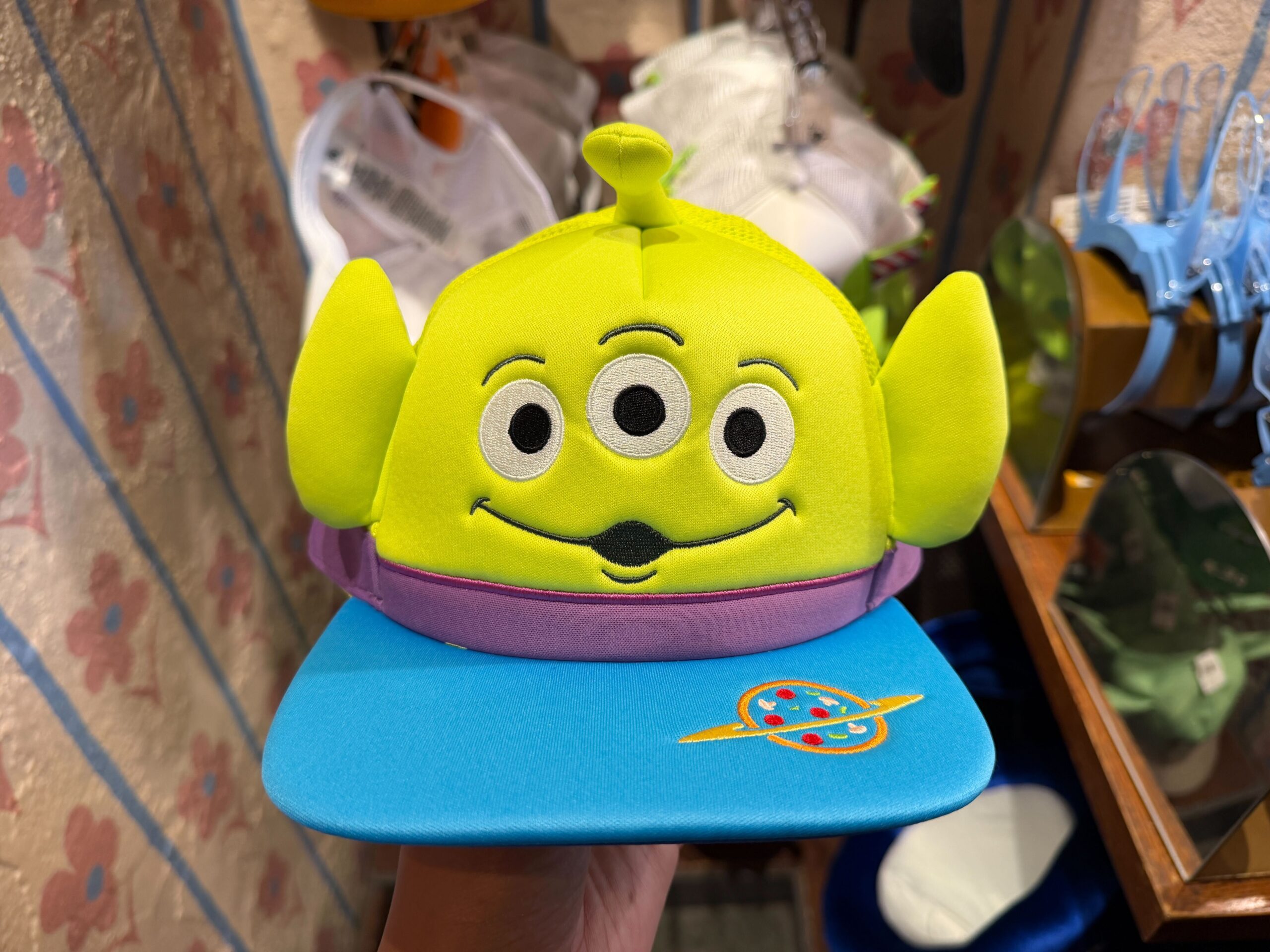 Channel the aliens from Toy Story with this cap. The top is green with large alien ears and embroidered details like eyes and a mouth. An antennae sticks out of the top. The band is purple while the flat bill is blue. It features an embroidered Pizza Planet logo.