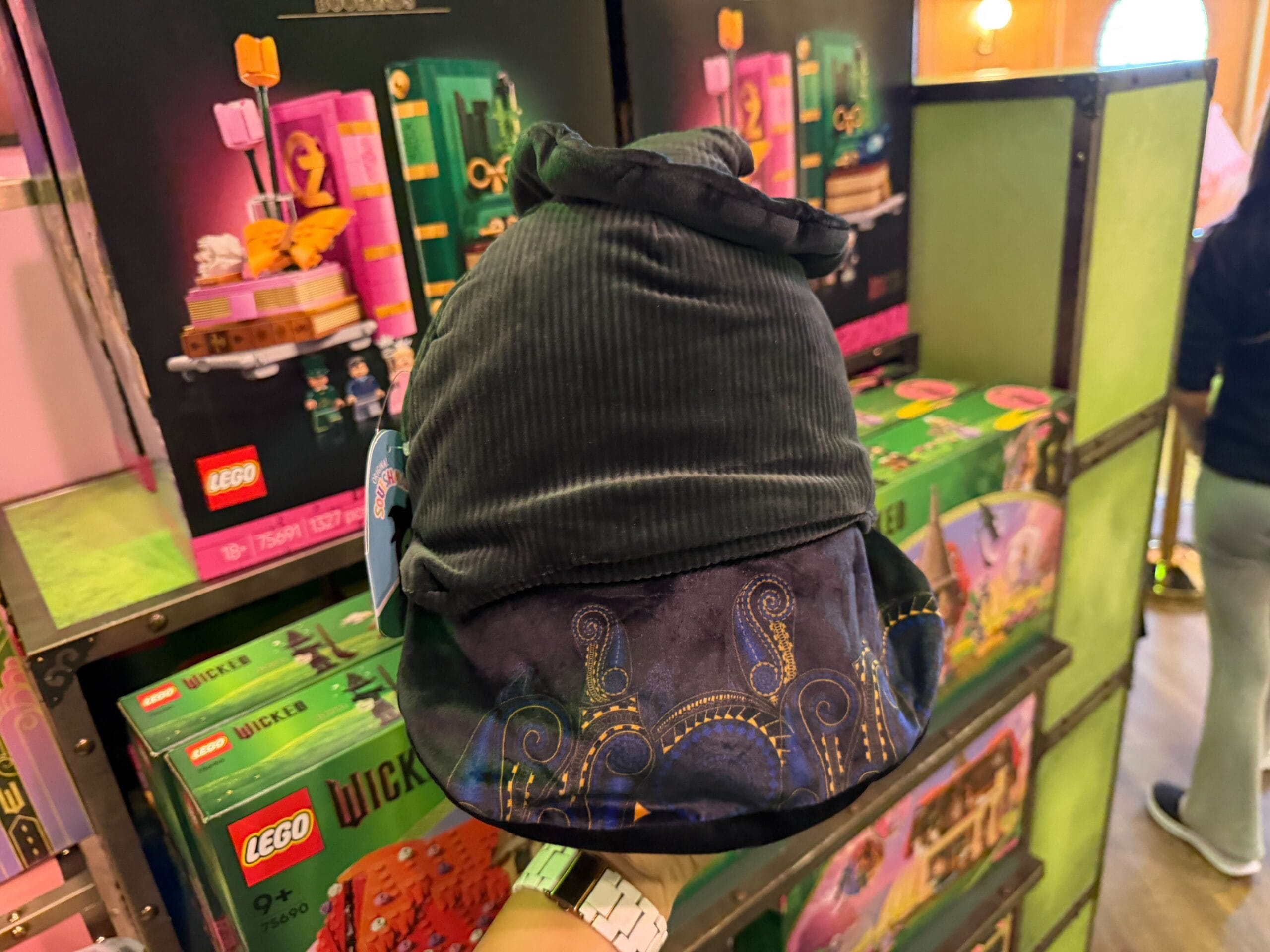 This Elphie Squishmallow shows the character wearing her black witch hat with a patterned cloak. Elphie's face details, including her freckles, are embroidered. Her face is green, and her cloak has yellow and purple pattern embellishments.